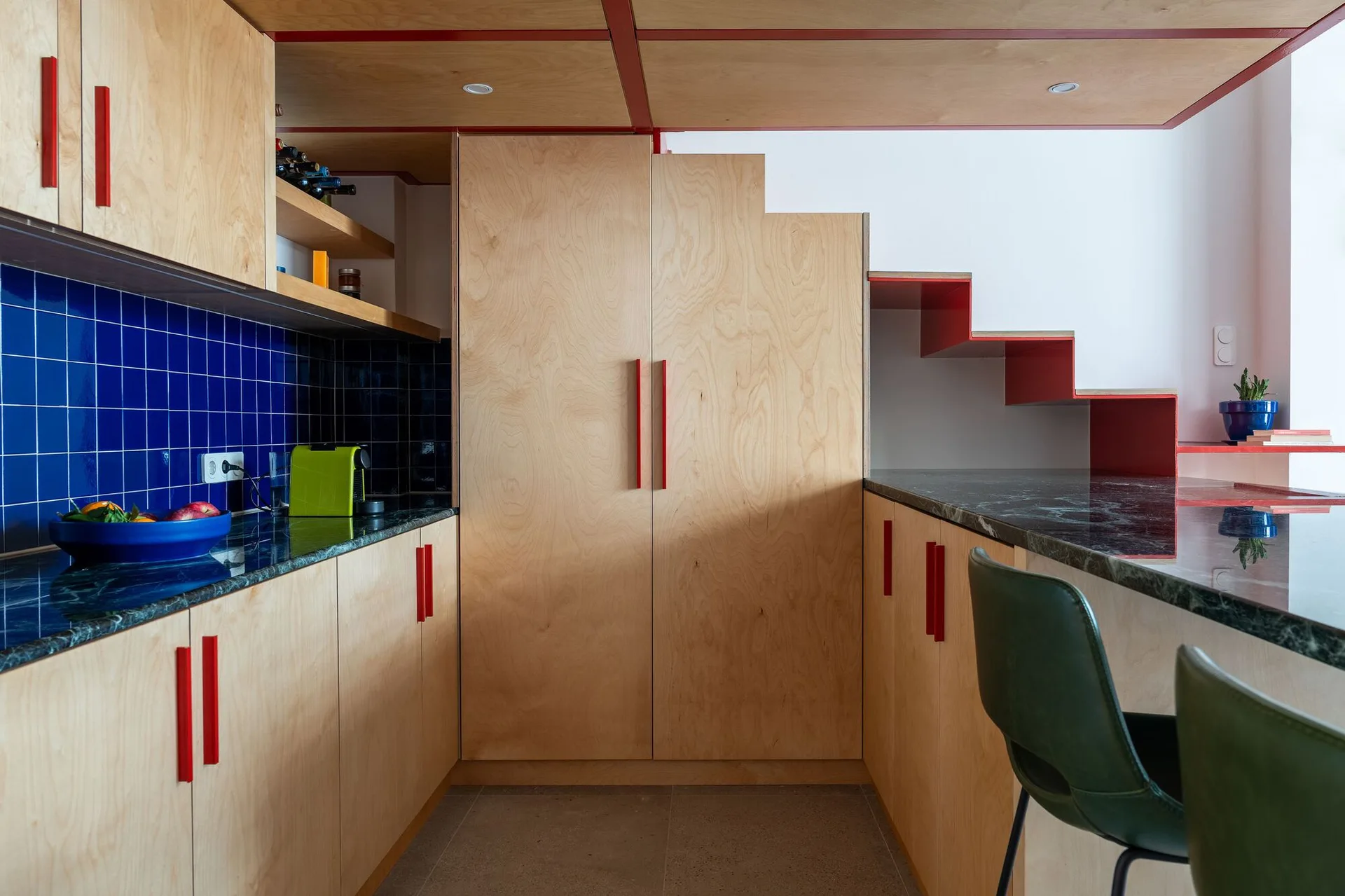 Modern kitchen featuring light plywood cabinets with red handles, a dark green countertop, blue tiled backsplash, and an integrated wooden staircase.