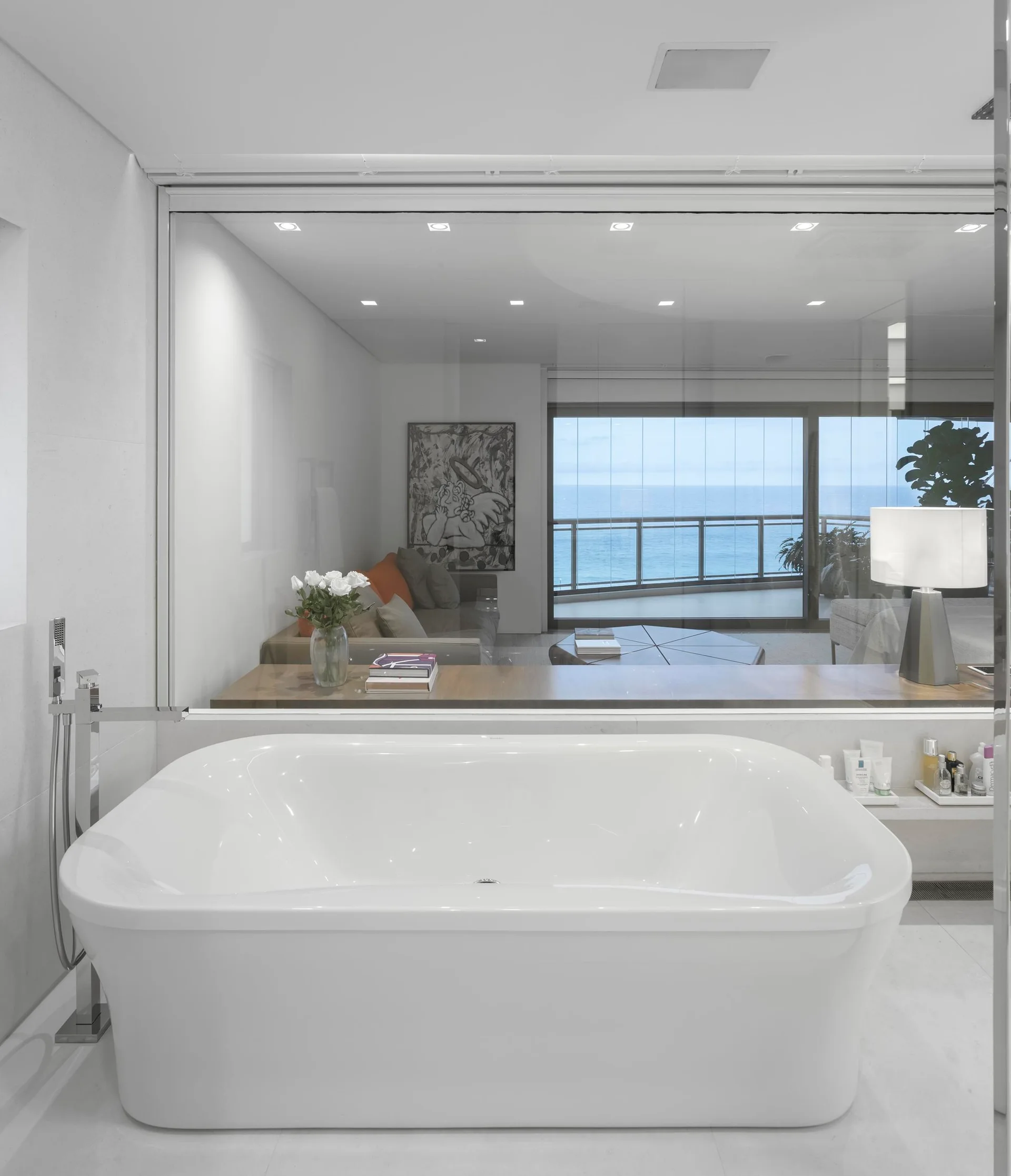 A modern bathroom with a white freestanding bathtub positioned before a large glass wall overlooking a living area and ocean view.