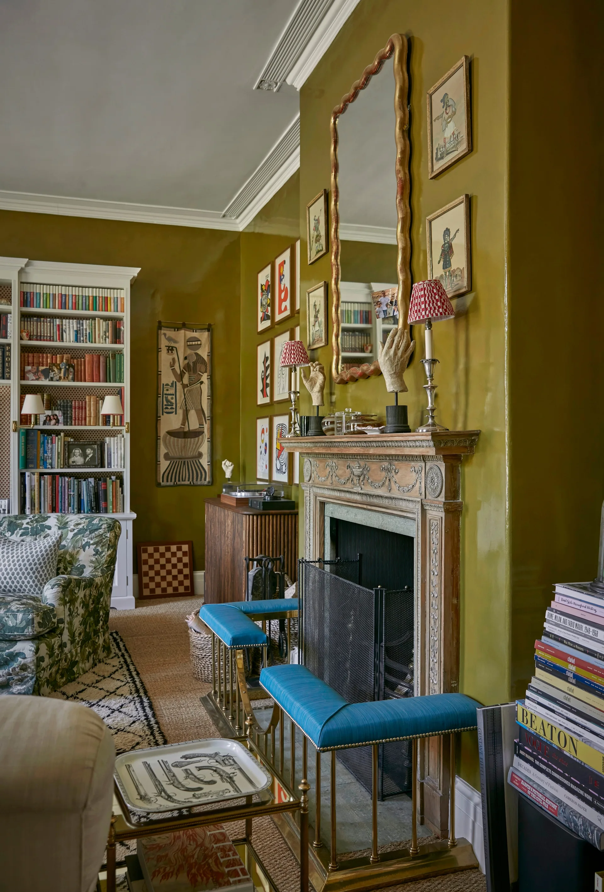 A traditional living room features glossy olive green walls, an ornate wood fireplace mantel with a scalloped gold mirror, and blue hearth stools.