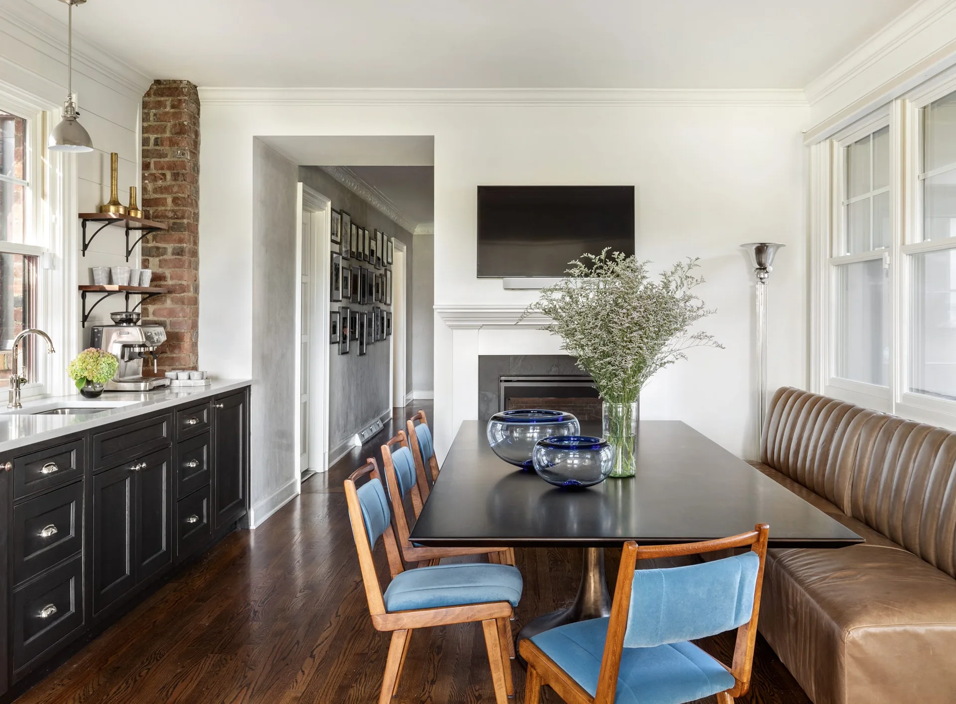 A dining room features a black table surrounded by three blue upholstered chairs and a long tufted brown leather banquette under windows.