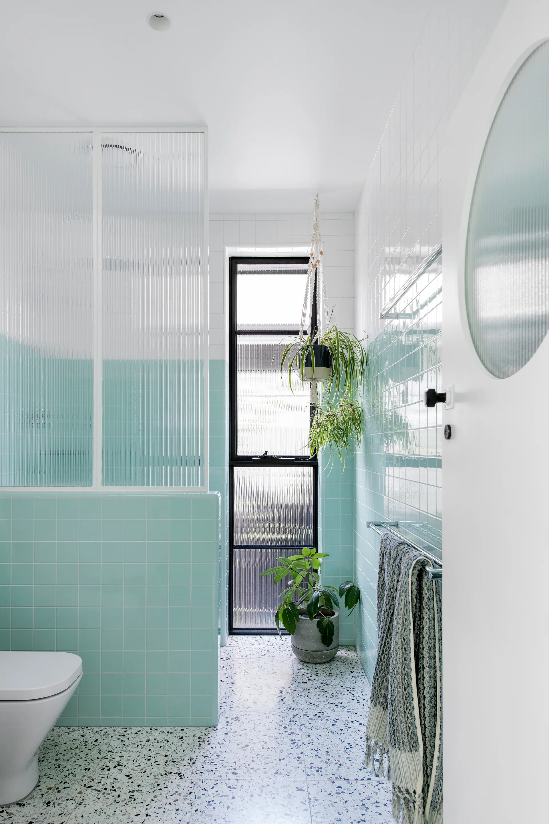 A modern bathroom features light teal wall tiles, terrazzo flooring, a black-framed window, and a fluted glass partition.