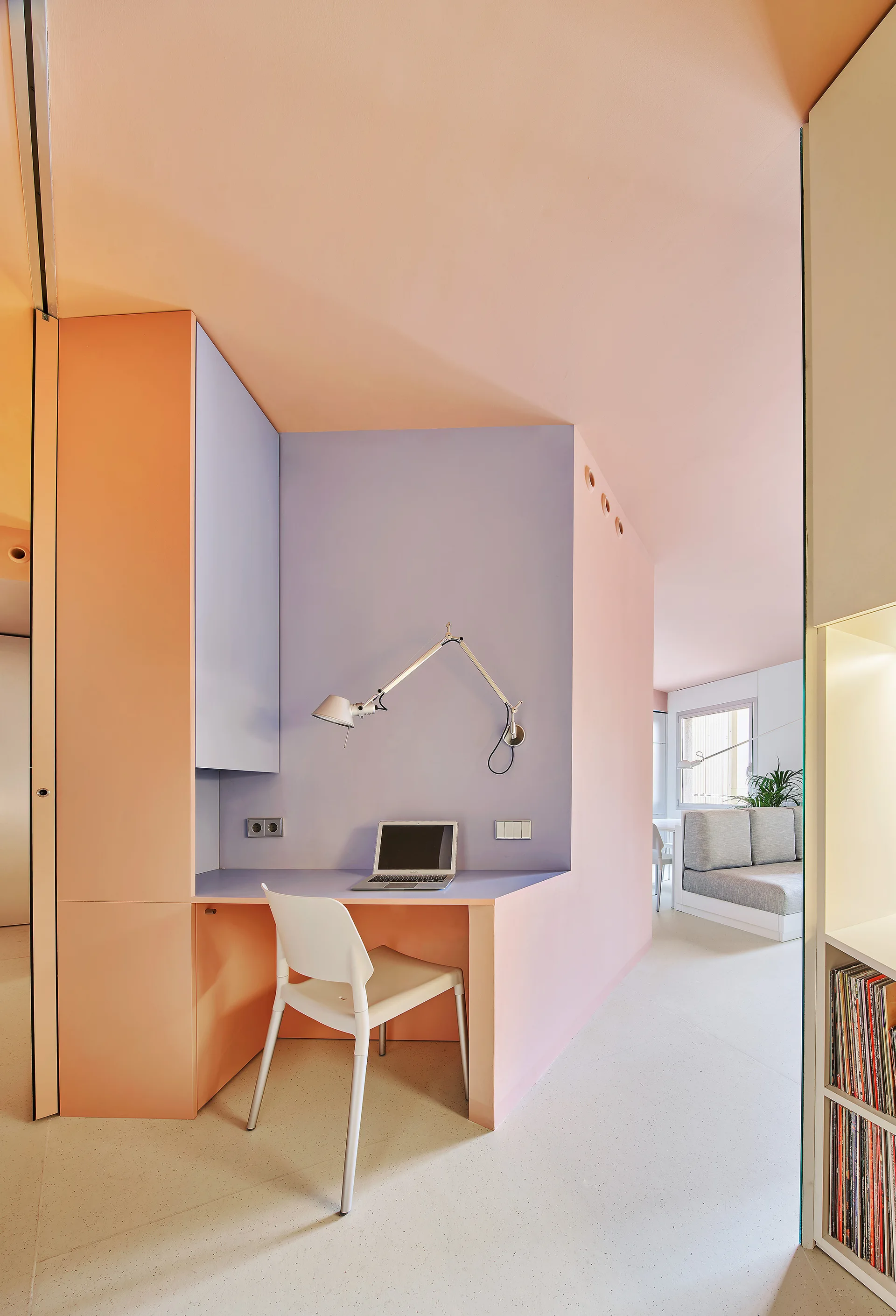 A modern home office features a built-in pastel peach and lavender desk with a white chair and an adjustable silver wall lamp.