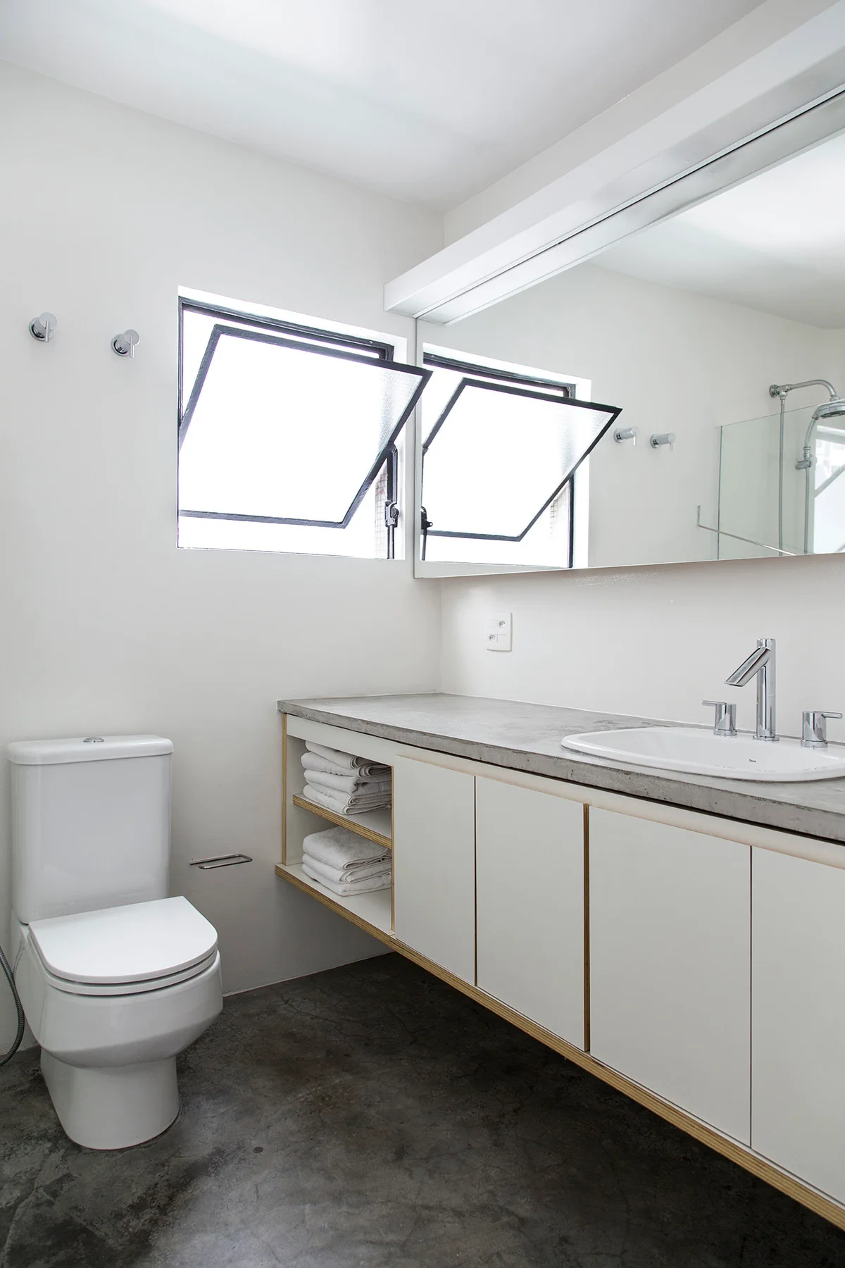 A minimalist bathroom features a white floating vanity with a concrete countertop, two open hopper windows, and a white toilet on a grey floor.