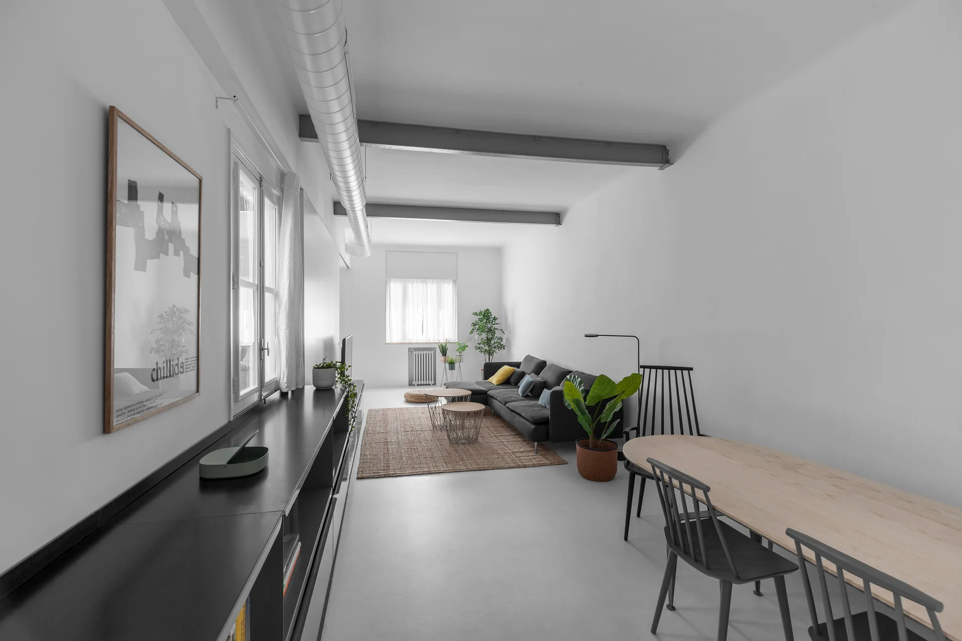 An industrial loft living space featuring a long black console, dark grey sectional sofa, and a light wood dining table under exposed ceiling ductwork.
