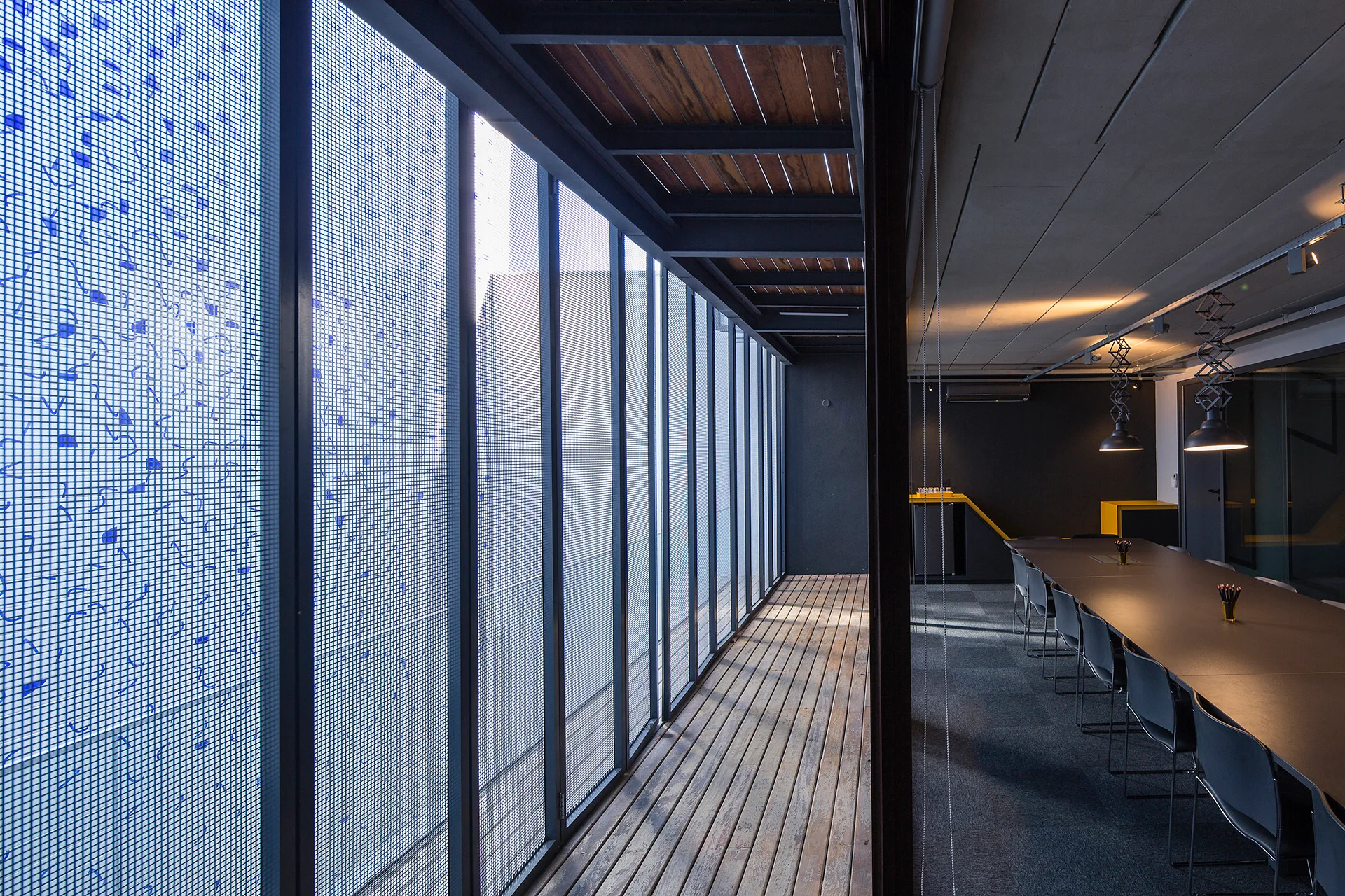 A modern corporate meeting room features a long dark conference table under pendant lights next to a glass corridor wall with a blue grid pattern.