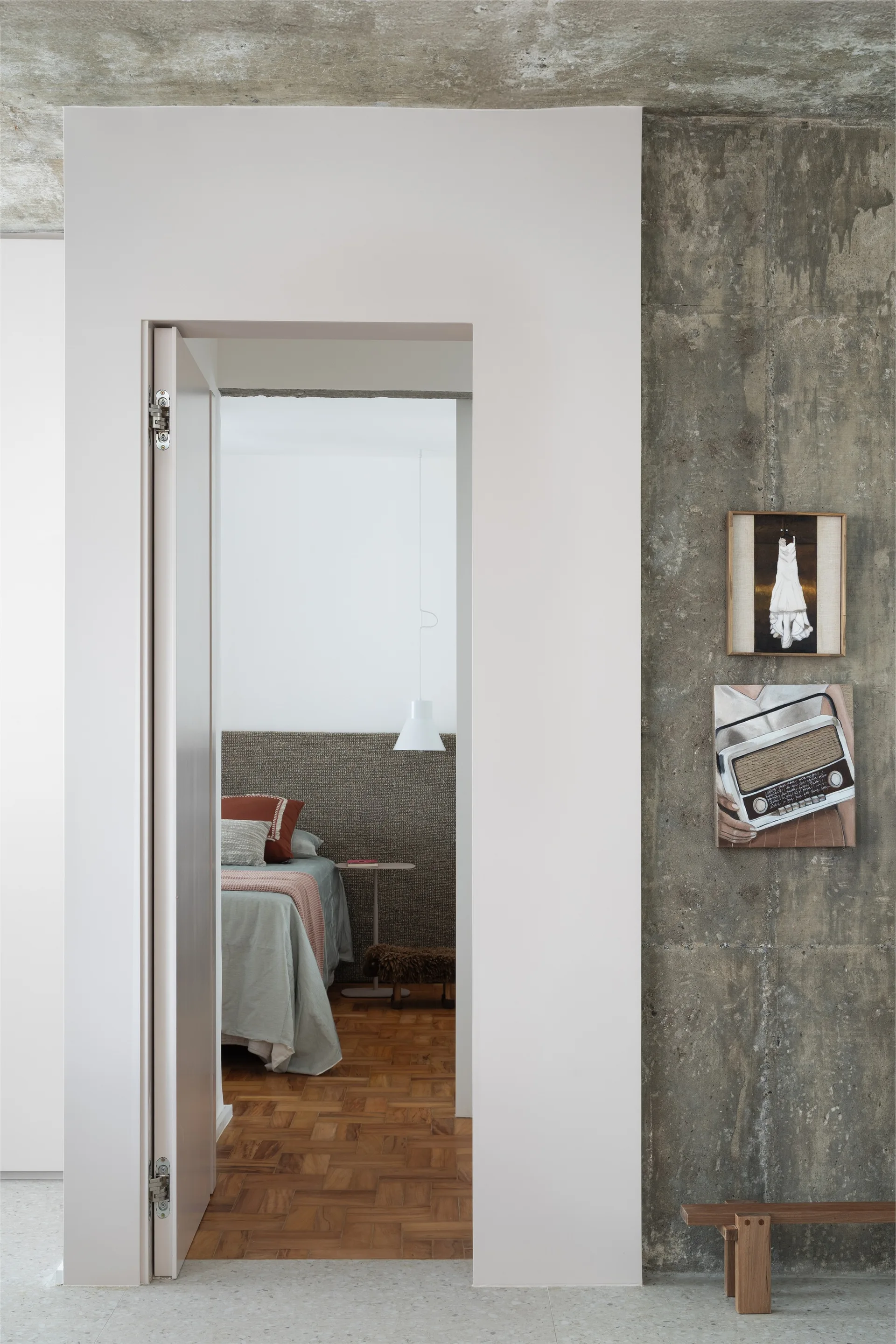 An open doorway reveals a bedroom with parquet wood floors and a woven headboard, situated beside a rough concrete wall featuring stacked artwork.