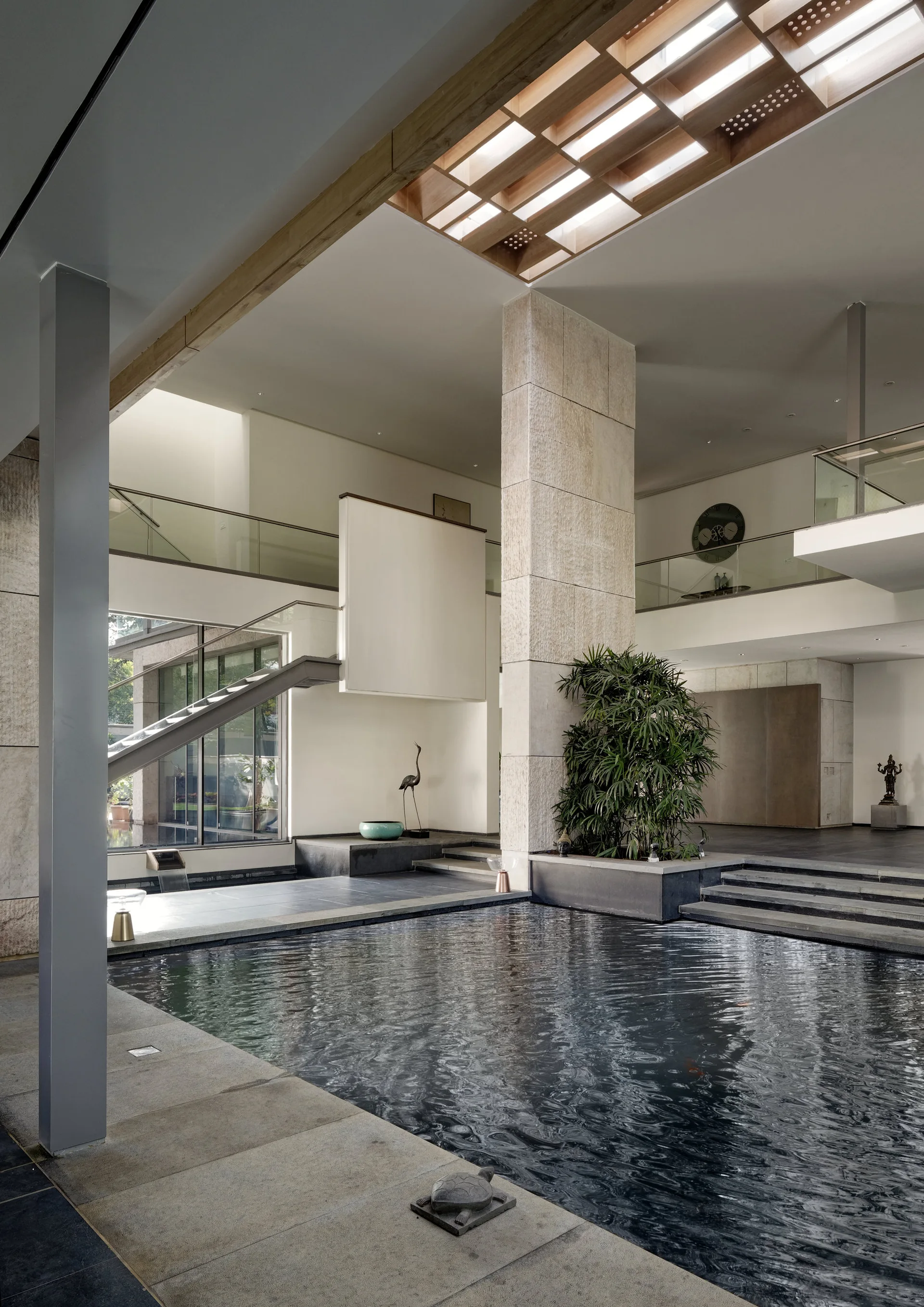 A modern indoor atrium features a dark reflection pool, a tall textured stone pillar, a grid skylight, and a mezzanine balcony.