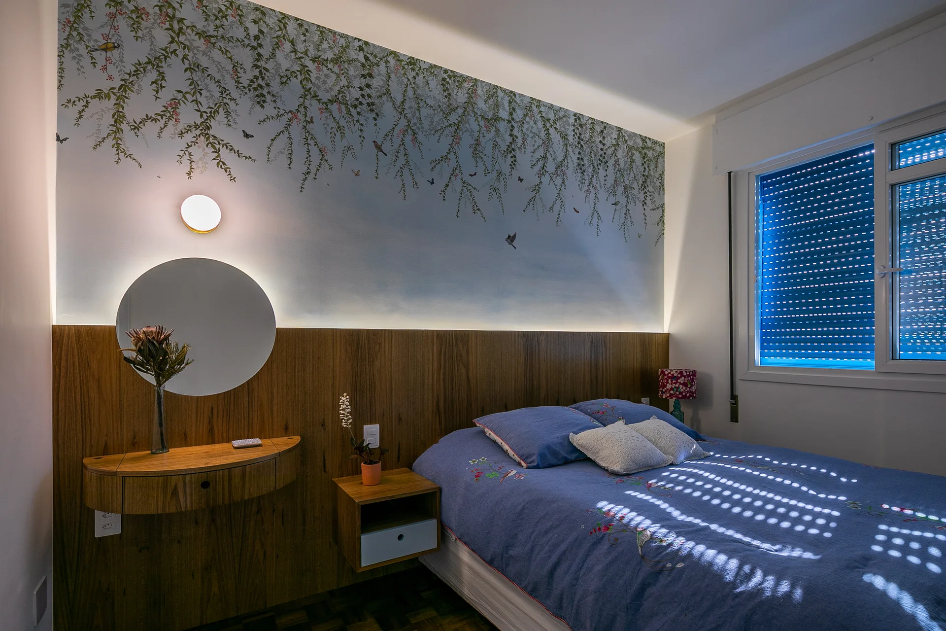 A contemporary bedroom featuring a blue bed next to wood-veneered wall paneling topped with a backlit botanical mural and a round mirror.