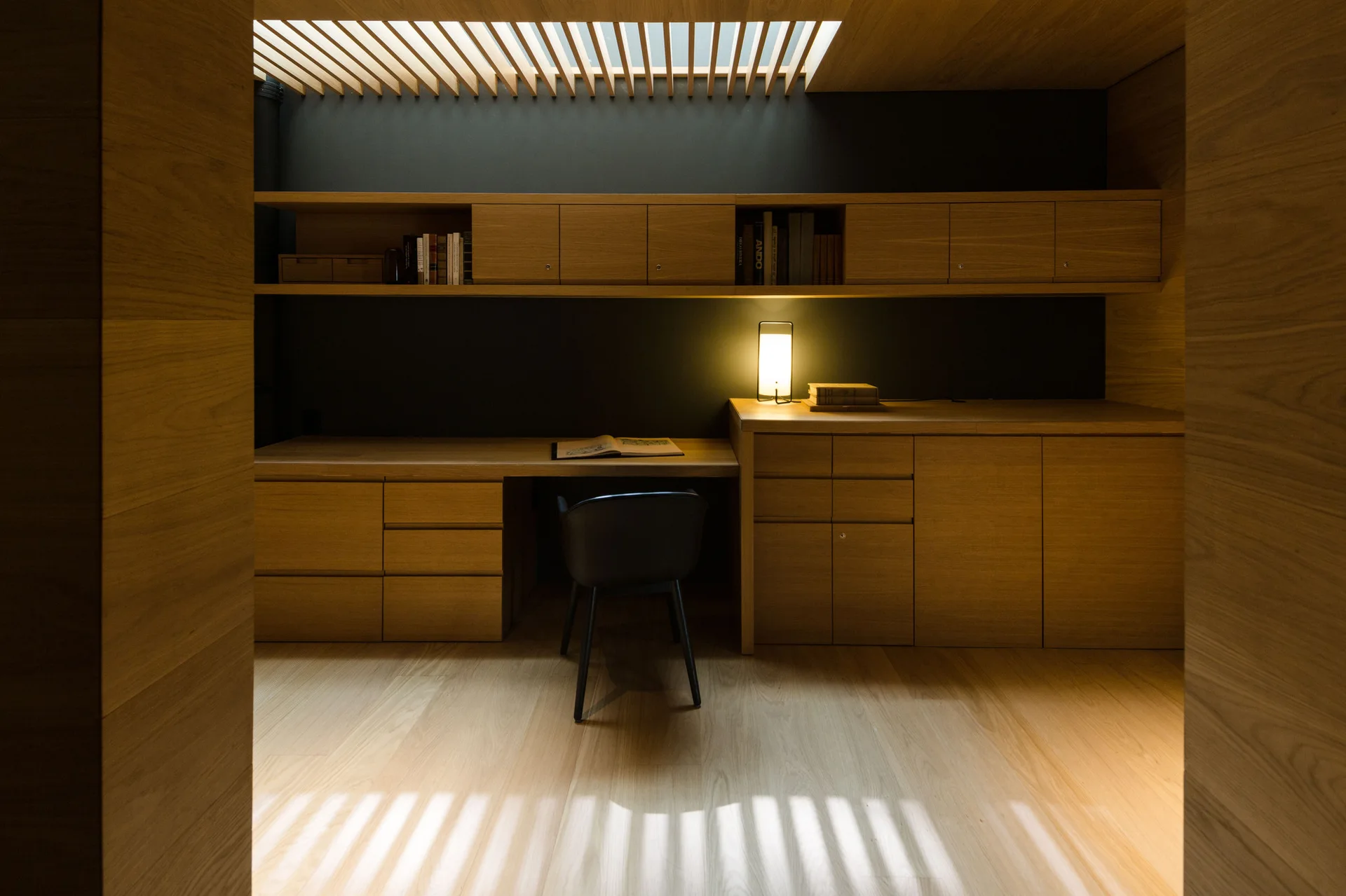A minimalist home office featuring a built-in wooden desk and matching shelves against a charcoal wall with a slatted skylight above.