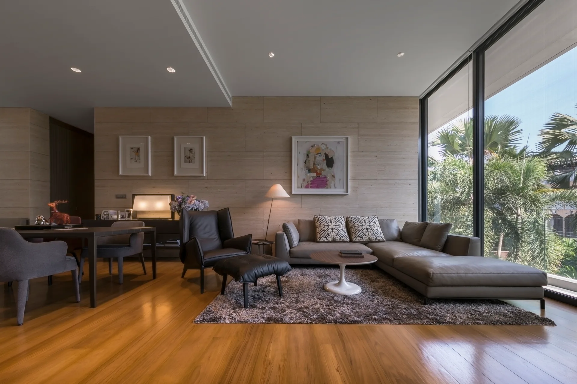 A contemporary living room featuring a grey leather sectional sofa, a stone tile accent wall, and floor-to-ceiling windows showing tropical trees.