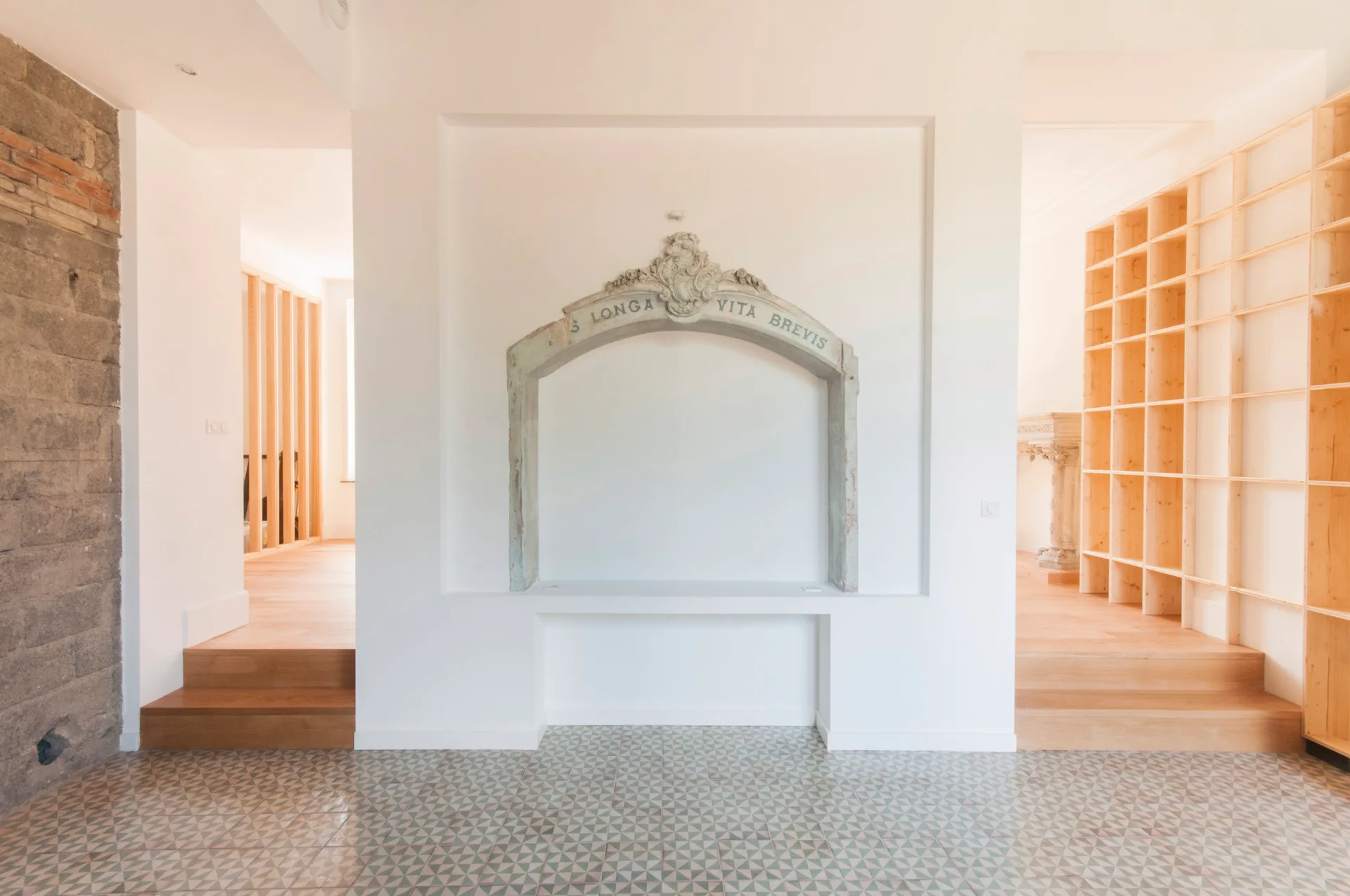 A bright room features an antique stone arch mounted inside a recessed white wall niche, alongside geometric tiled floors and light wood steps.