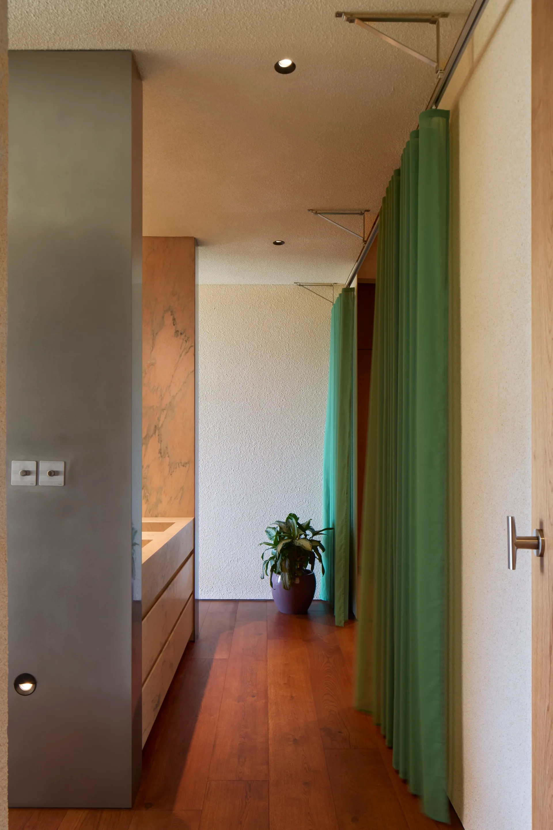 A modern bathroom hallway features rich wooden floors, textured cream walls, and a ceiling-mounted green fabric curtain used as a space divider.