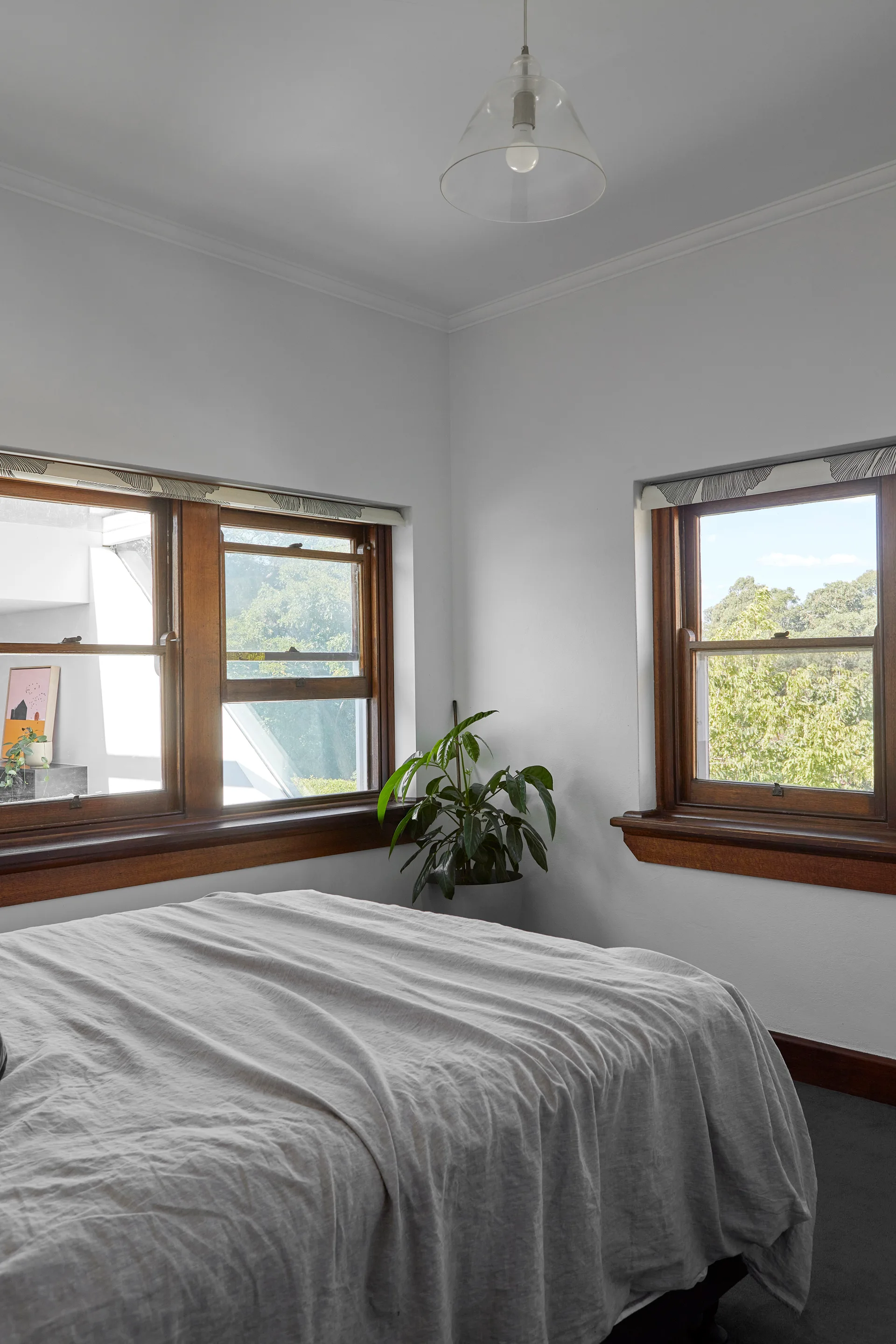 A minimalist bedroom features a bed with grey linen bedding, two warm wood-framed windows, white walls, and a potted corner plant.