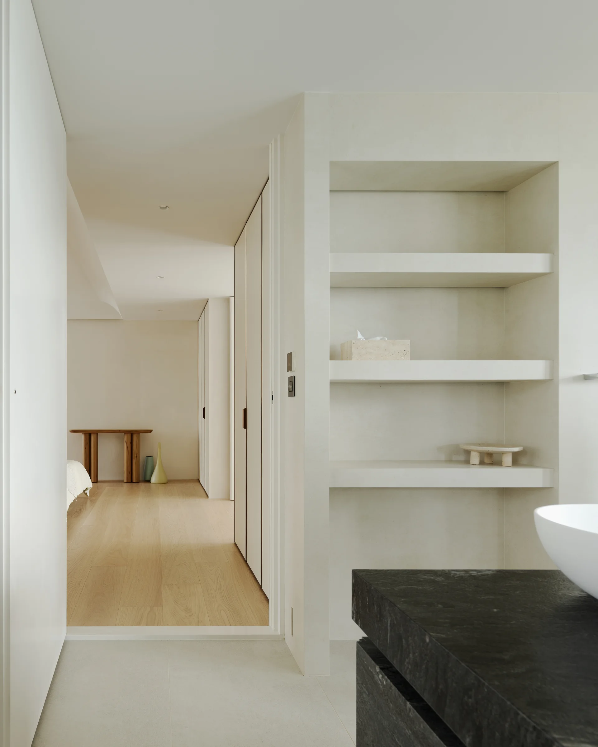 A minimalist bathroom features a dark stone vanity with a white vessel sink next to a recessed plaster shelving unit looking into a light-flooded hallway.