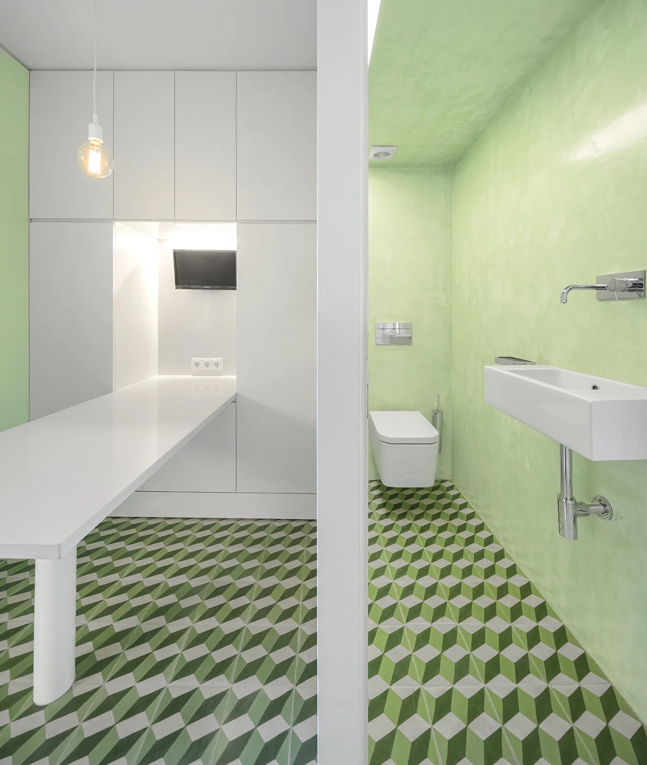 A split view of a minimalist bathroom with green plaster walls and a white workspace area, both sharing a green and white geometric tiled floor.