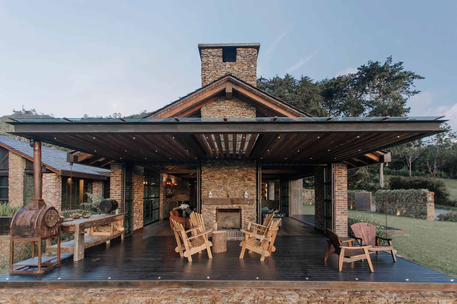 A covered outdoor patio features a central stone fireplace, dark wood plank flooring, and rustic wooden lounge chairs under a wood-beamed roof.