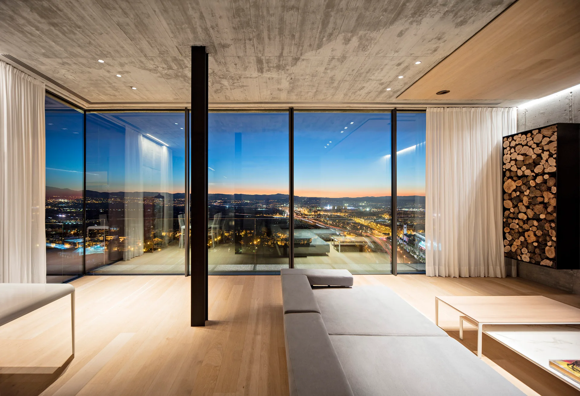 A minimalist living room with a low grey sofa, light wood floors, a concrete ceiling, and floor-to-ceiling windows showing a city sunset.