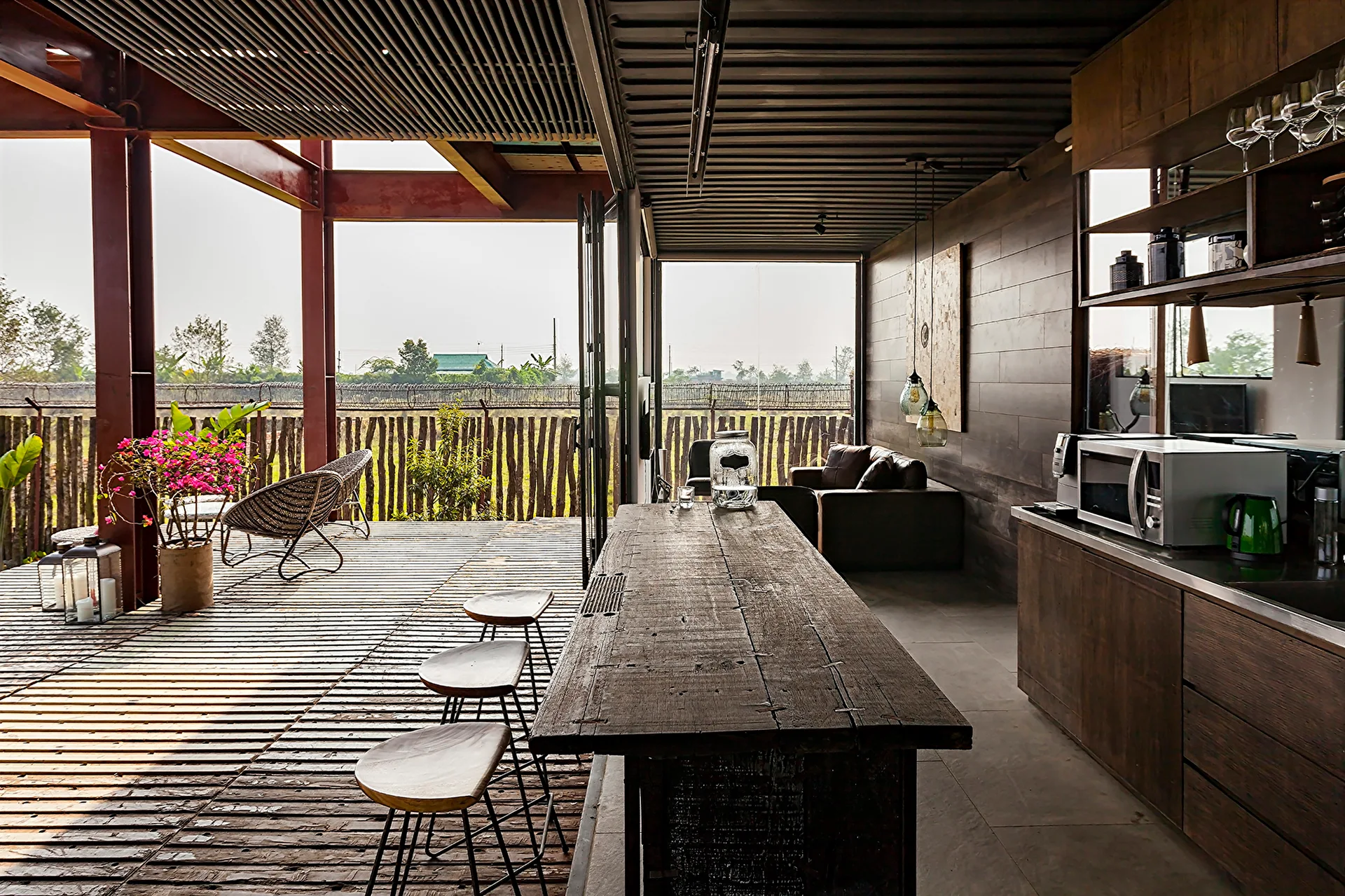 An industrial kitchen built inside a shipping container features a weathered wood island and opens onto a spacious slatted wood deck with outdoor seating.