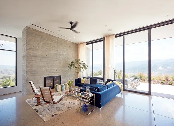 A modern living room with a textured grey concrete wall, a navy blue velvet sectional sofa, and expansive glass sliding doors showing a mountain view.