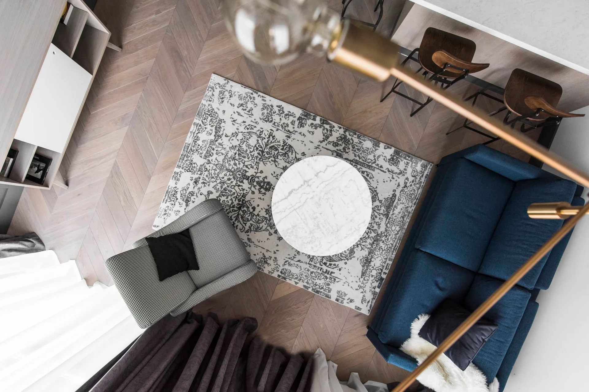 A high-angle view of a living room featuring a blue sofa, a houndstooth armchair, a round marble coffee table, and herringbone wood flooring.