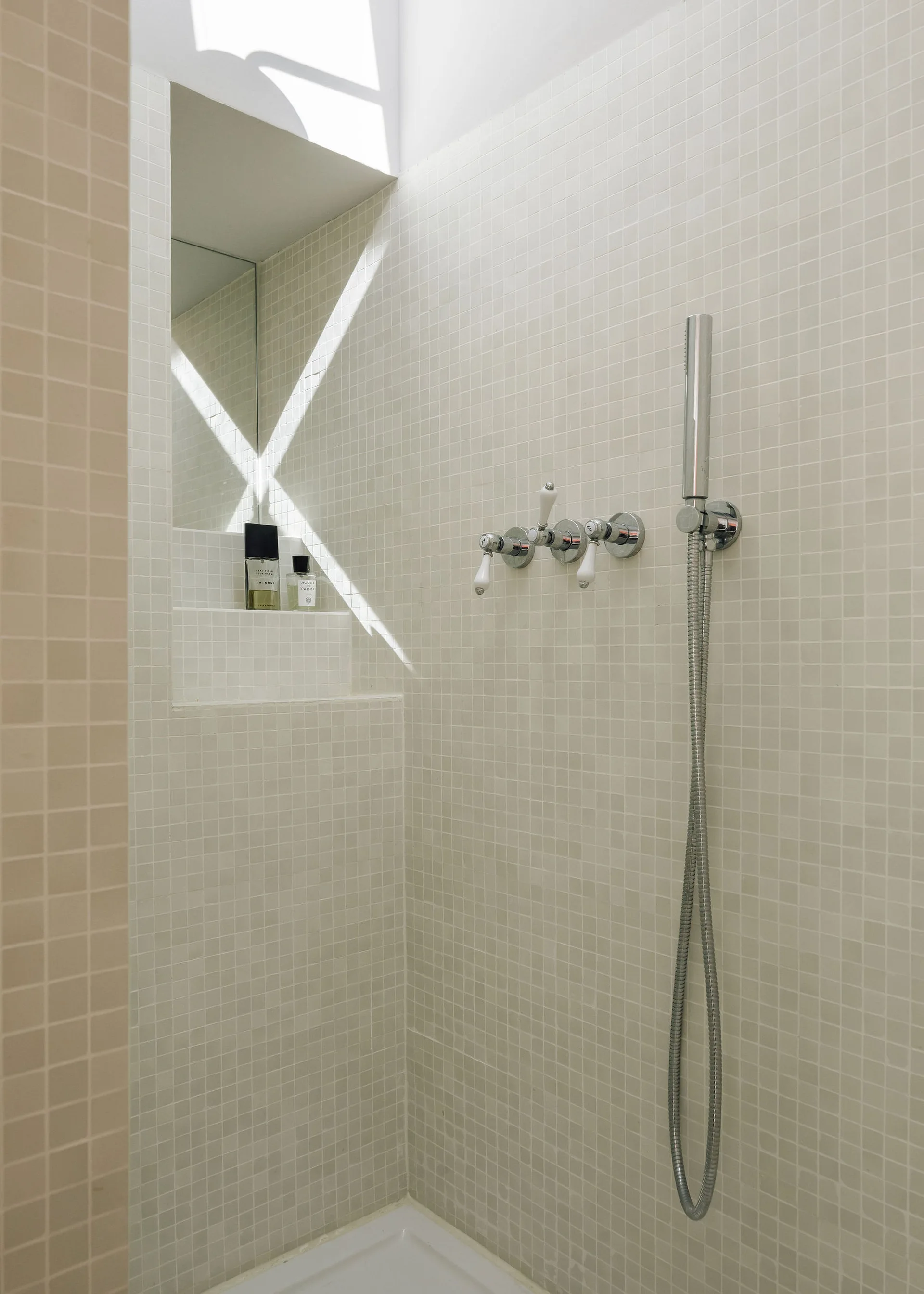 A shower stall features off-white mosaic tiled walls, chrome cross-handle faucets, a handheld shower head, and a deep recessed niche with a mirror.