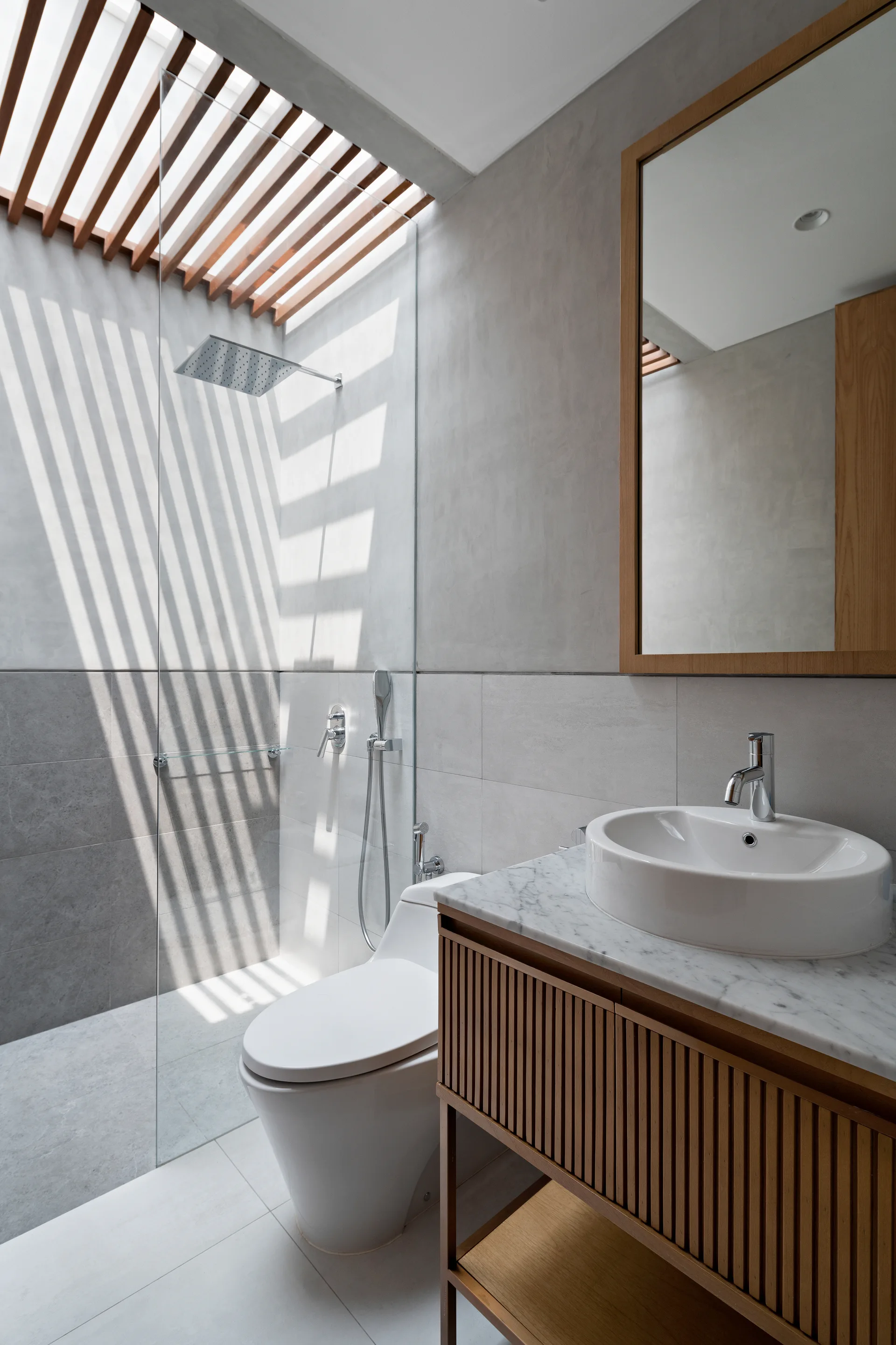 Gray tiled bathroom featuring a walk-in glass shower under a wooden slatted skylight, a white toilet, and a fluted wood vanity with a marble top.