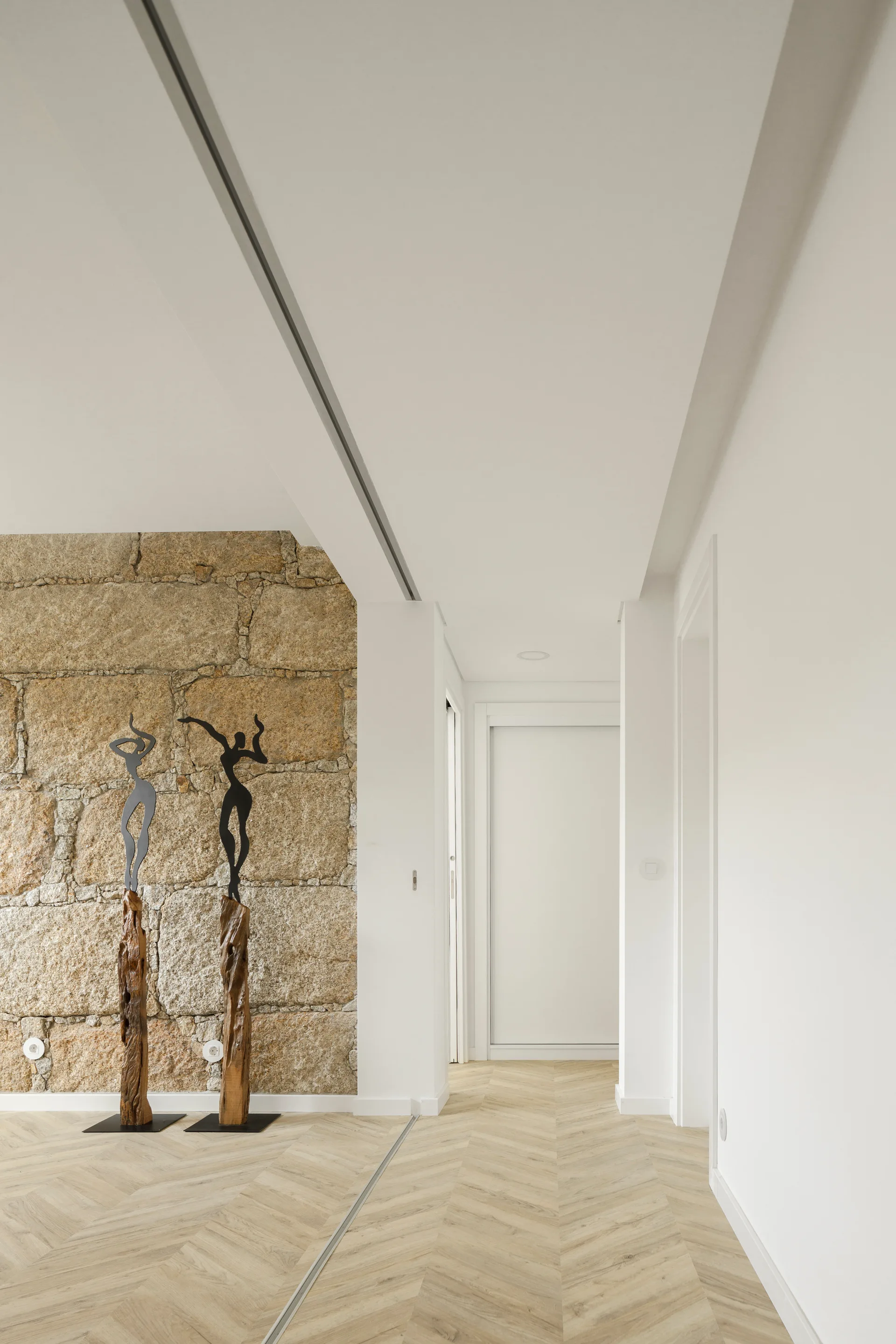 A minimalist white hallway features light wood herringbone floors, a textured exposed stone wall, and two abstract black metal sculptures.