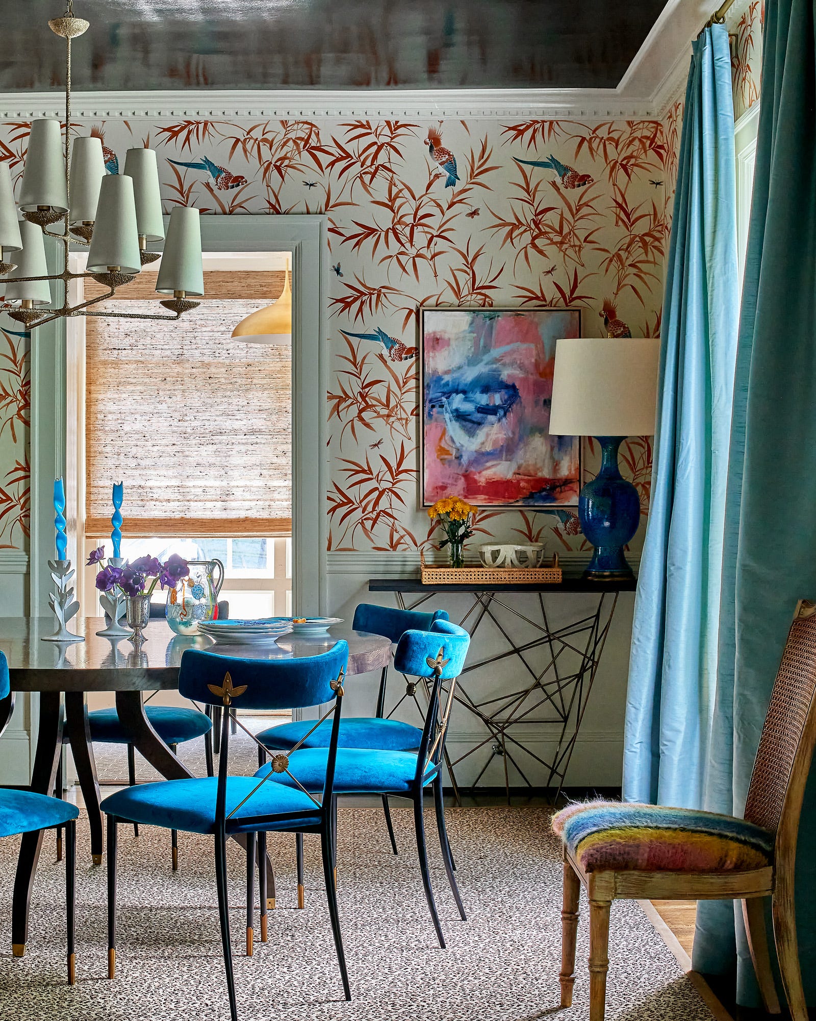 Blue velvet chairs surround a round dining table in a room featuring bamboo patterned wallpaper, blue curtains, and an animal print rug.