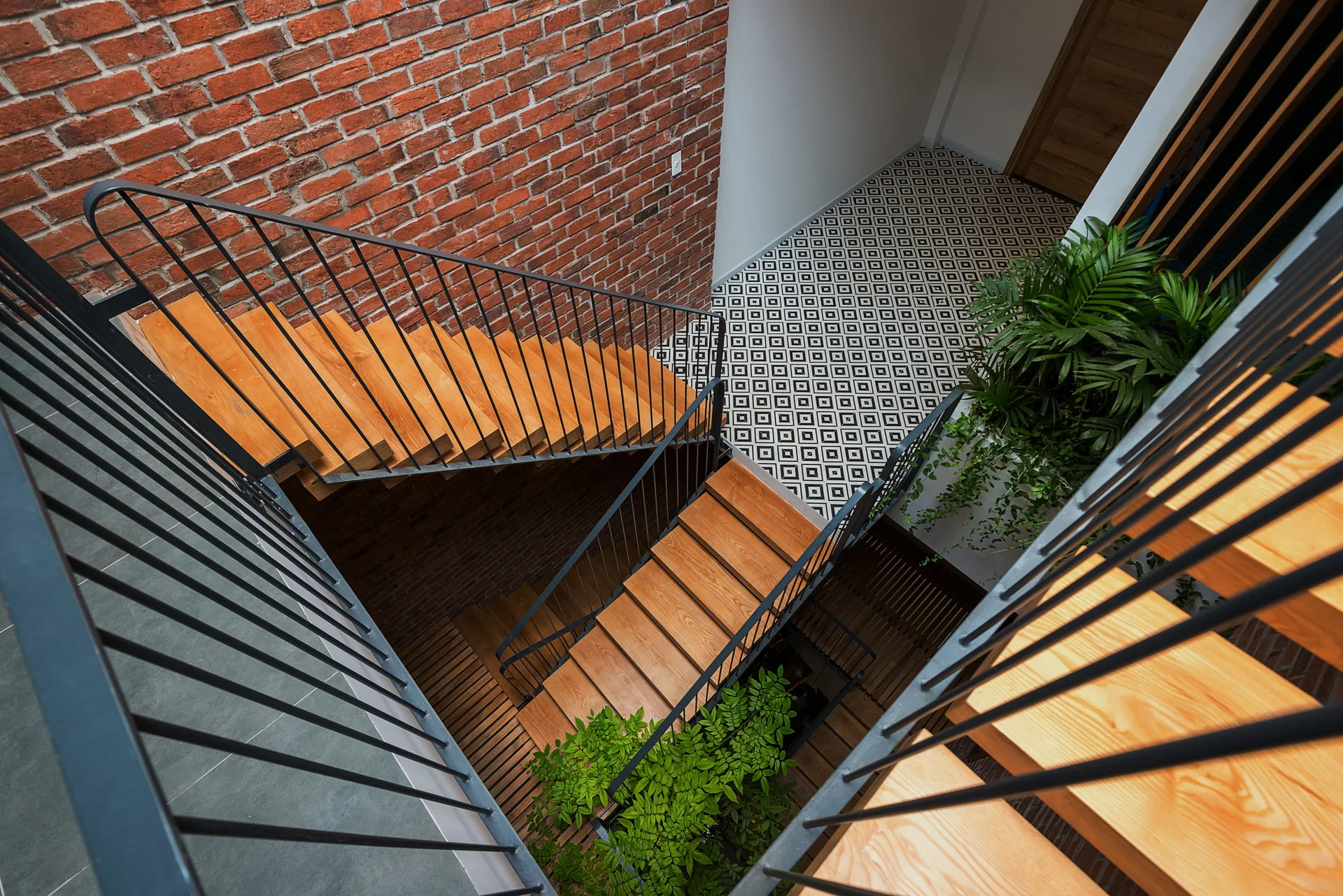 A top-down view of a wooden staircase with black metal railings next to an exposed red brick wall and black-and-white geometric tiled floor.