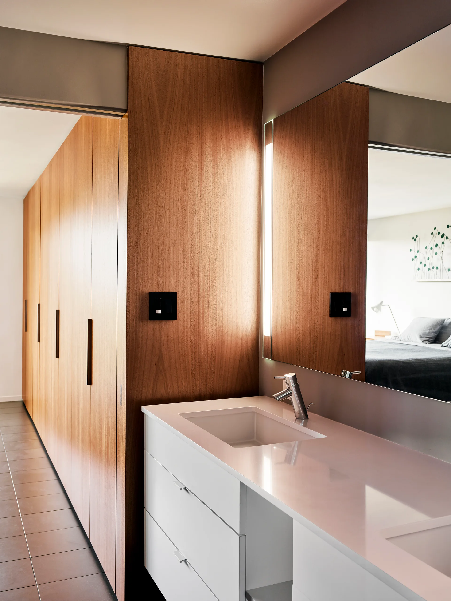A white bathroom vanity with a chrome faucet stands next to floor-to-ceiling wood closets and a large wall mirror.