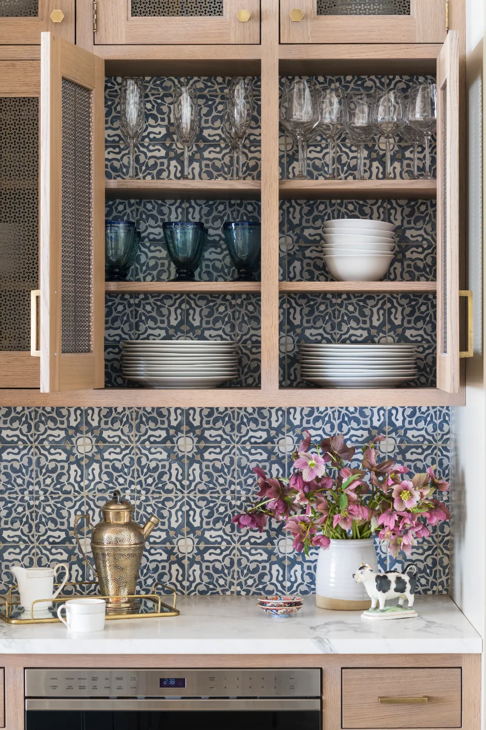 Open light oak cabinets display white plates and glassware against a blue and white patterned tile backsplash on a marble countertop.