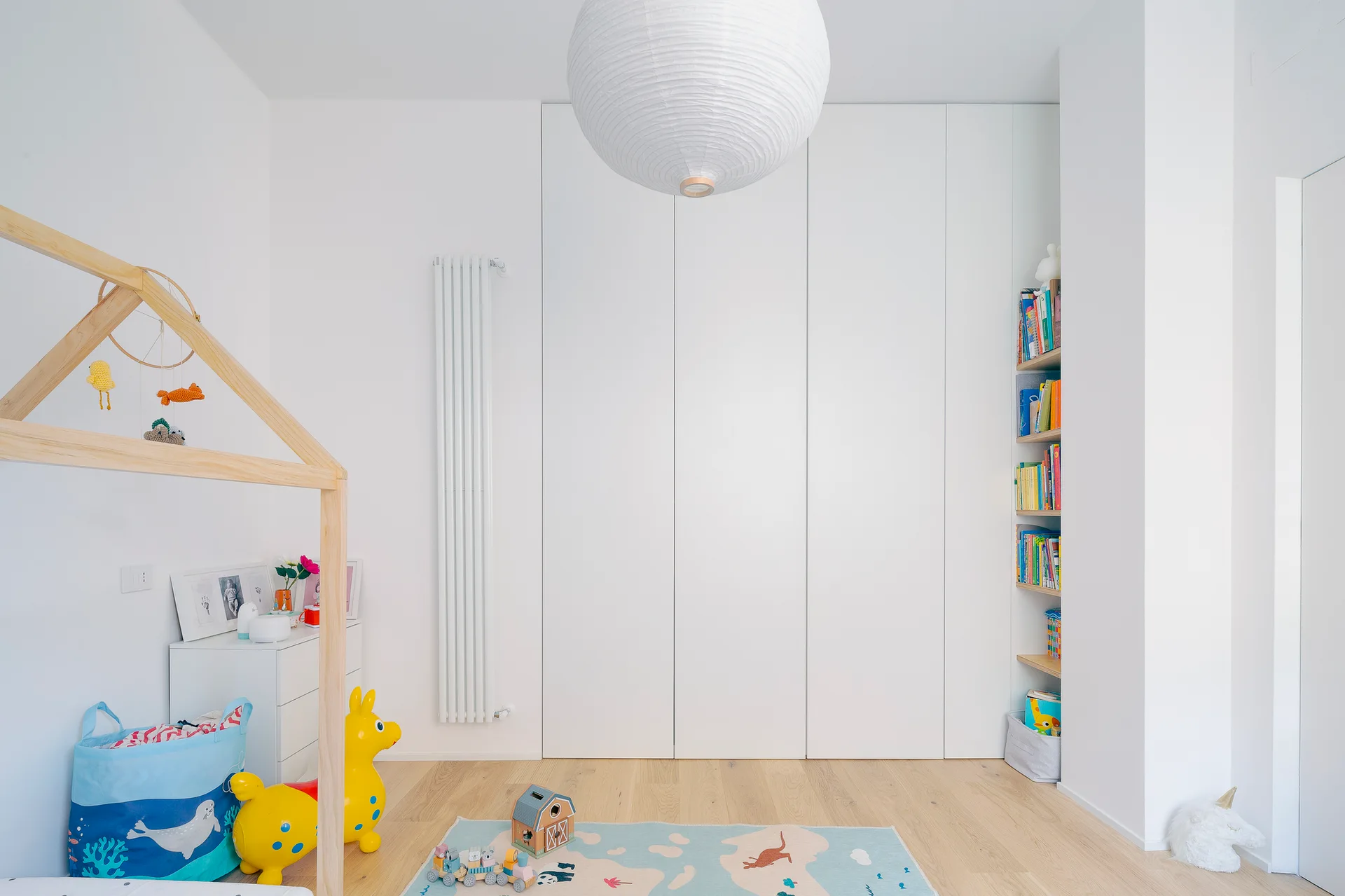 A bright kids' room features a wooden house-frame bed, flat-panel white wardrobes, a vertical radiator, and a large paper globe pendant light.
