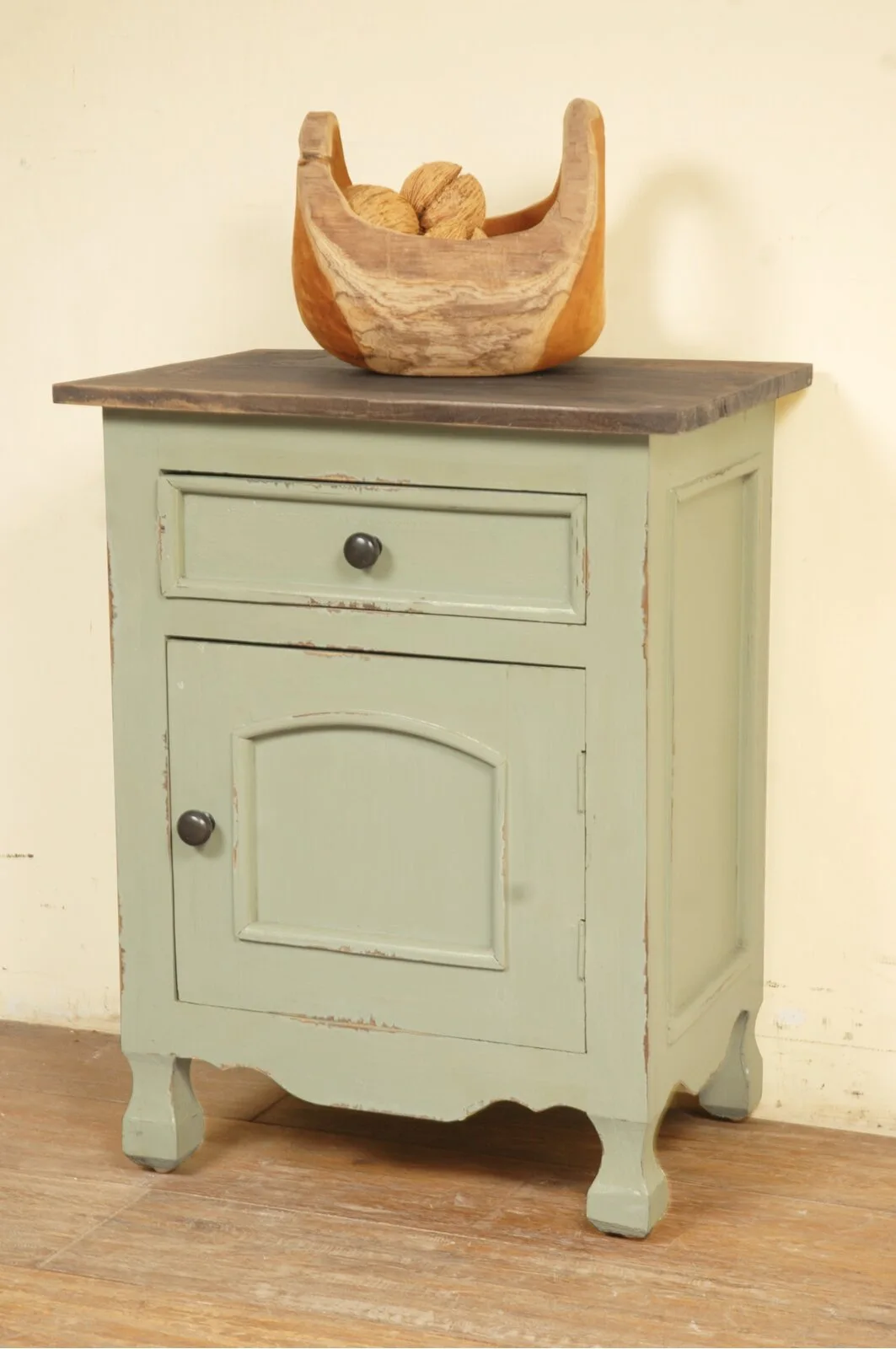 A distressed sage green wooden nightstand with a dark brown top features a carved wood bowl filled with decorative nuts.