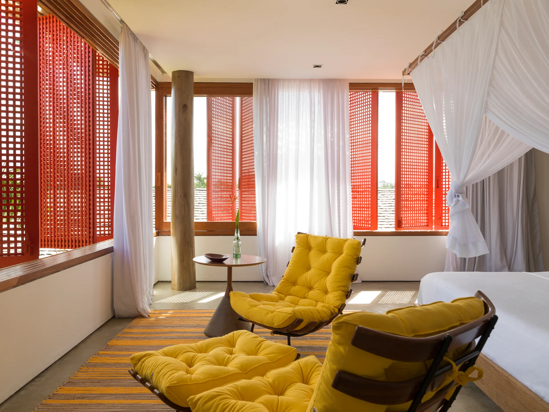 A bright bedroom features two tufted yellow lounge chairs, a canopy bed with white fabric, and bright red grid-patterned window shutters.