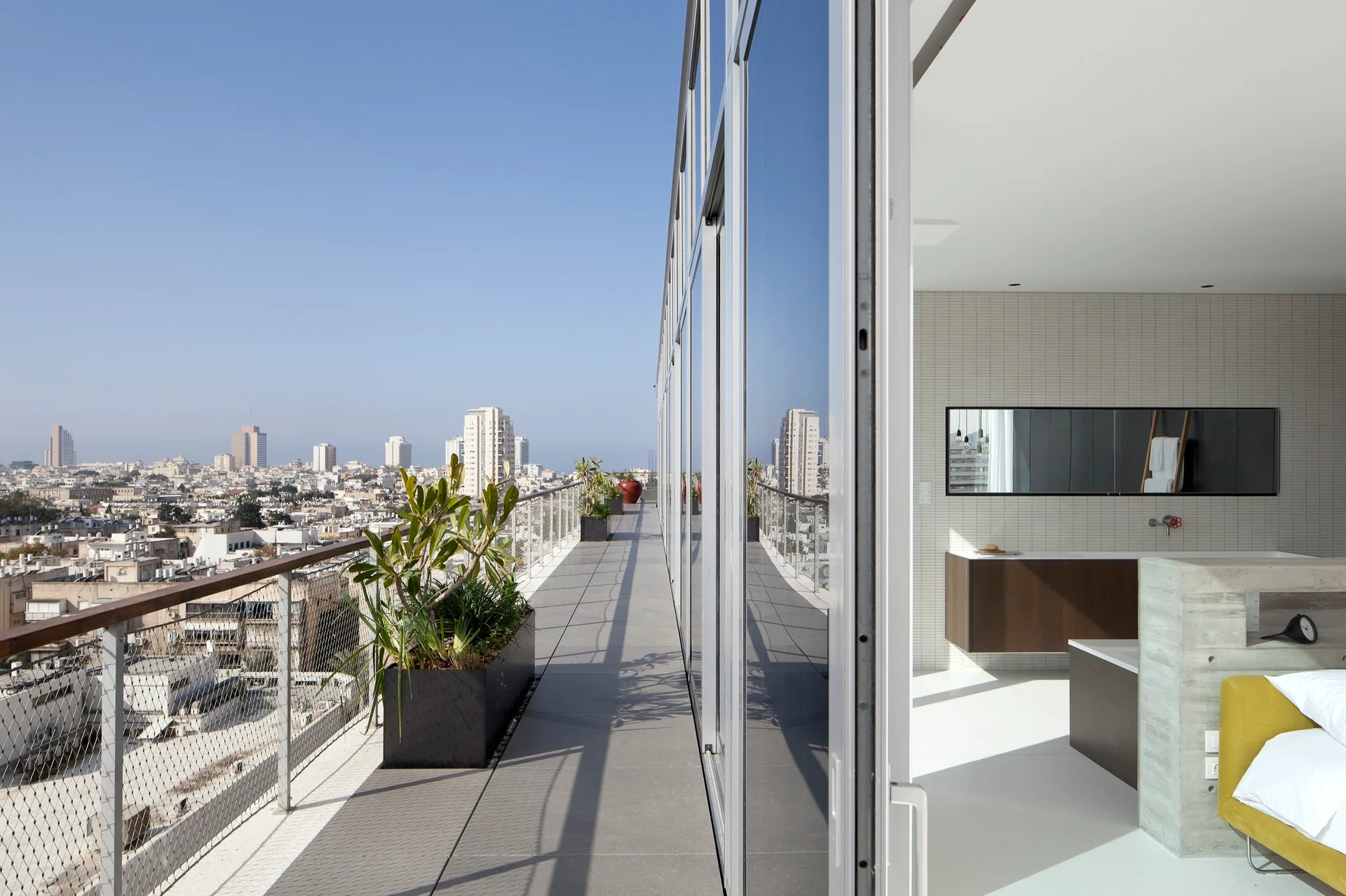 A long high-rise balcony with metal railings and black planters features sliding glass doors that open to a modern bedroom and bathroom suite.