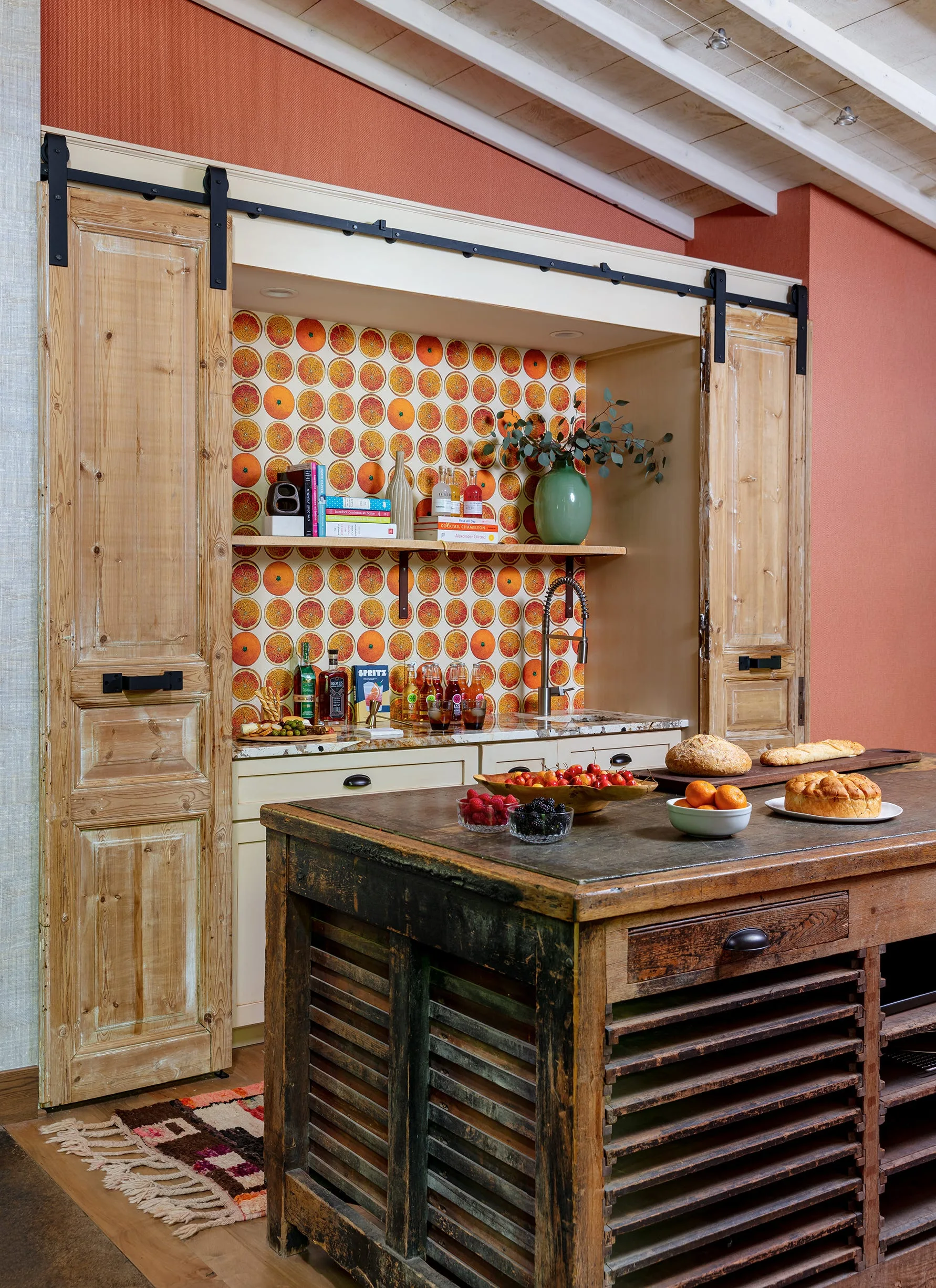 A rustic home bar features a recessed niche with orange citrus wallpaper, sliding barn doors, and a weathered wood island in the foreground.