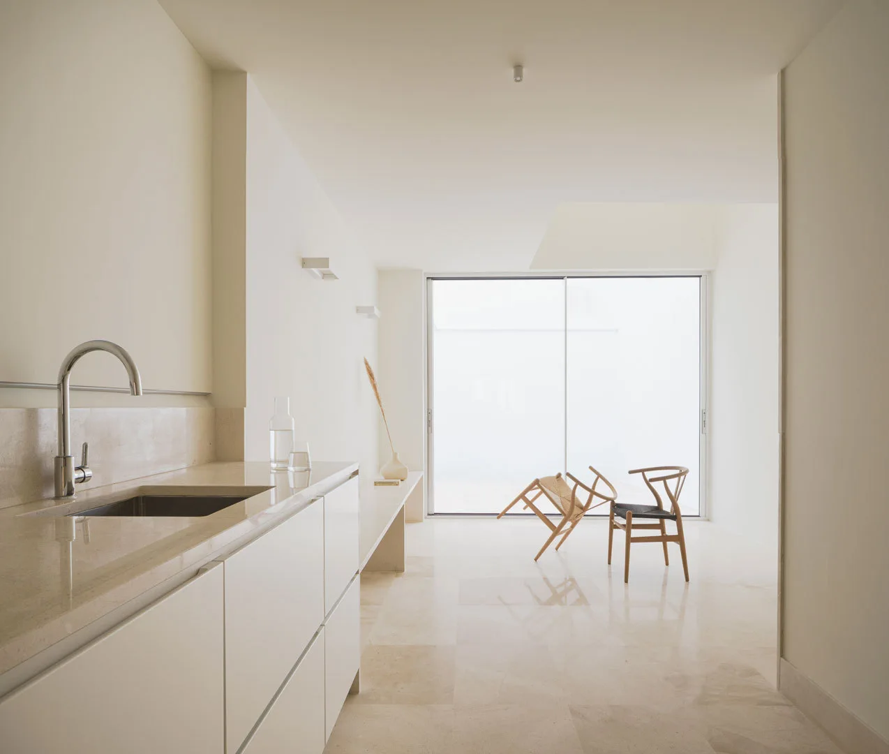 A minimalist off-white kitchen featuring sleek white cabinets, beige stone tile flooring, and two wooden chairs near a large sliding glass door.