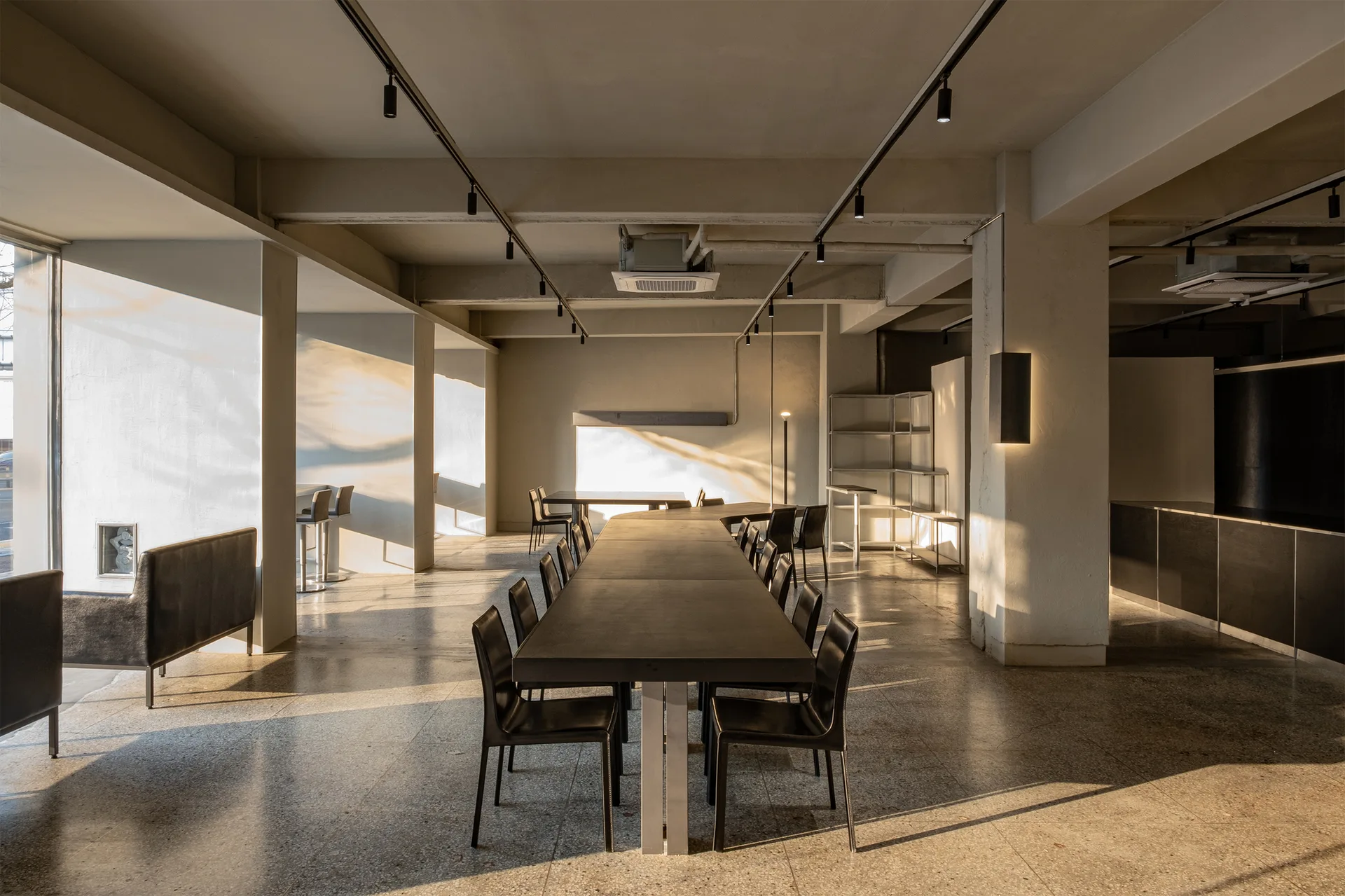 A long dark table with black chairs sits in a spacious room featuring polished concrete floors, exposed ceiling beams, and warm sunlight.