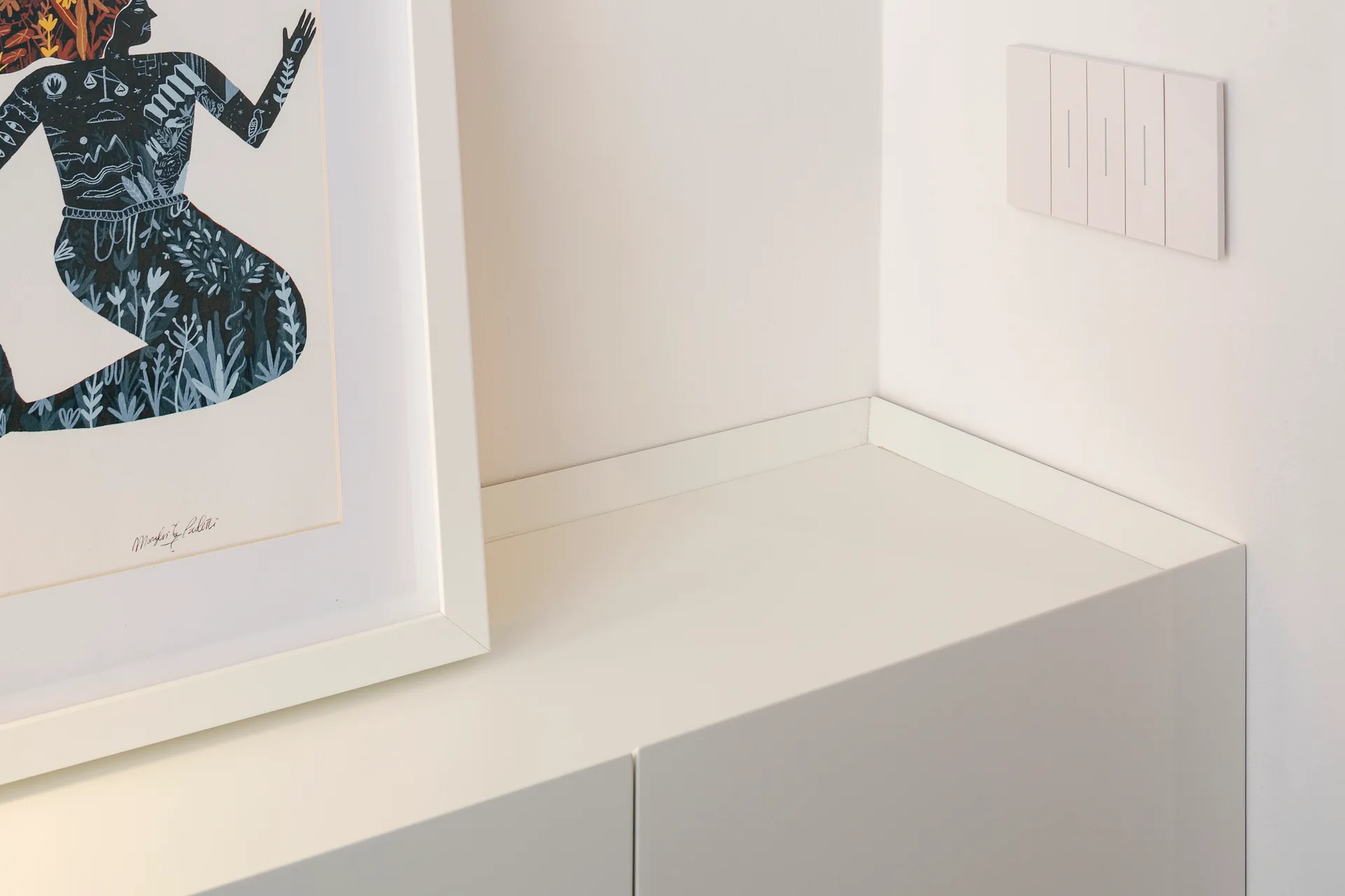 A close-up of a matte white cabinet in a corner featuring a white multi-gang light switch on the wall and a leaning framed print.