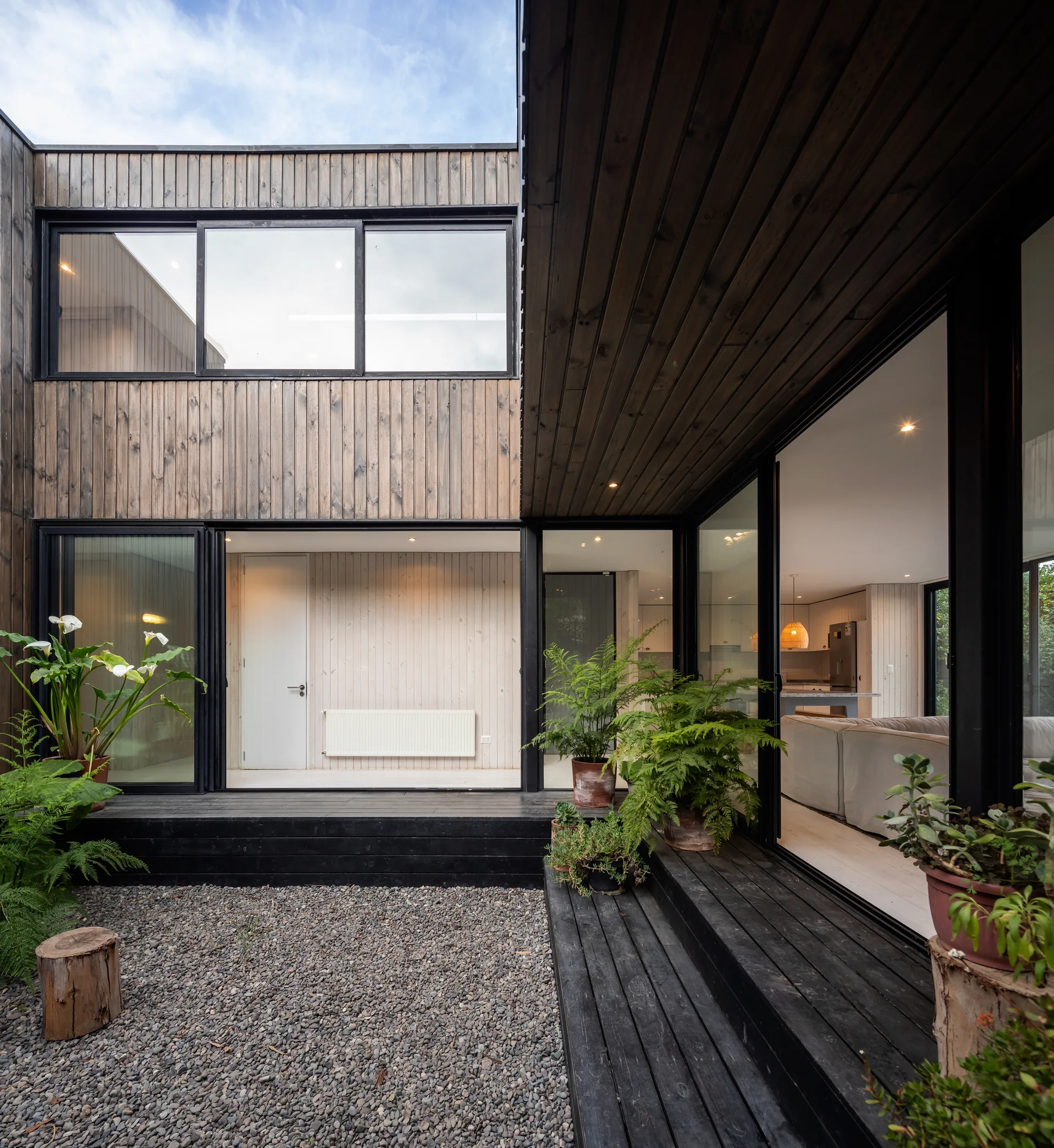 A modern enclosed courtyard features a black-stained wooden deck, gravel ground, vertical timber siding, and large glass sliding doors.