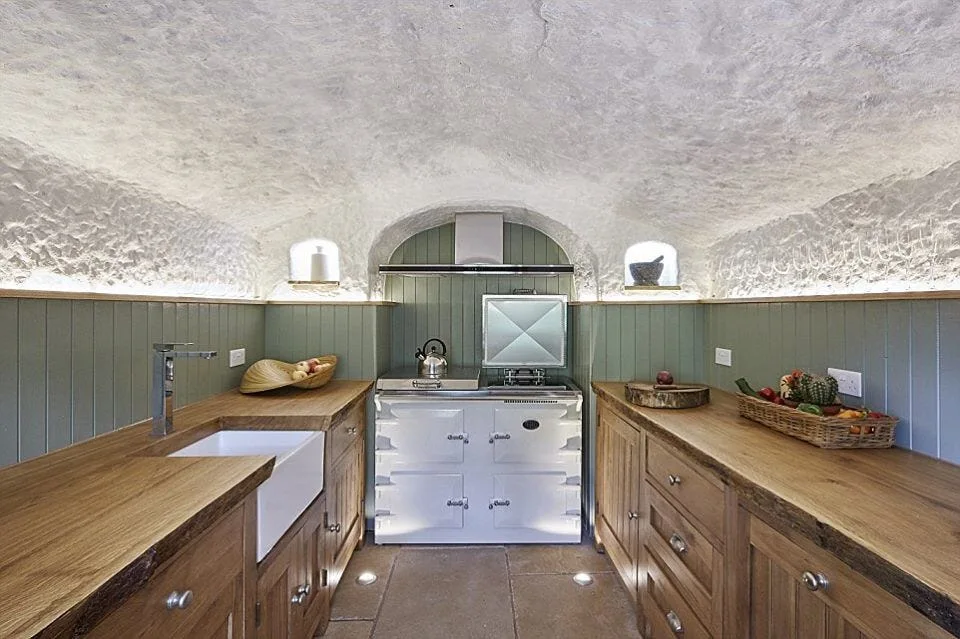 A rustic kitchen features a curved textured stone ceiling above sage green vertical wood paneling, wood cabinets, and a white range cooker.