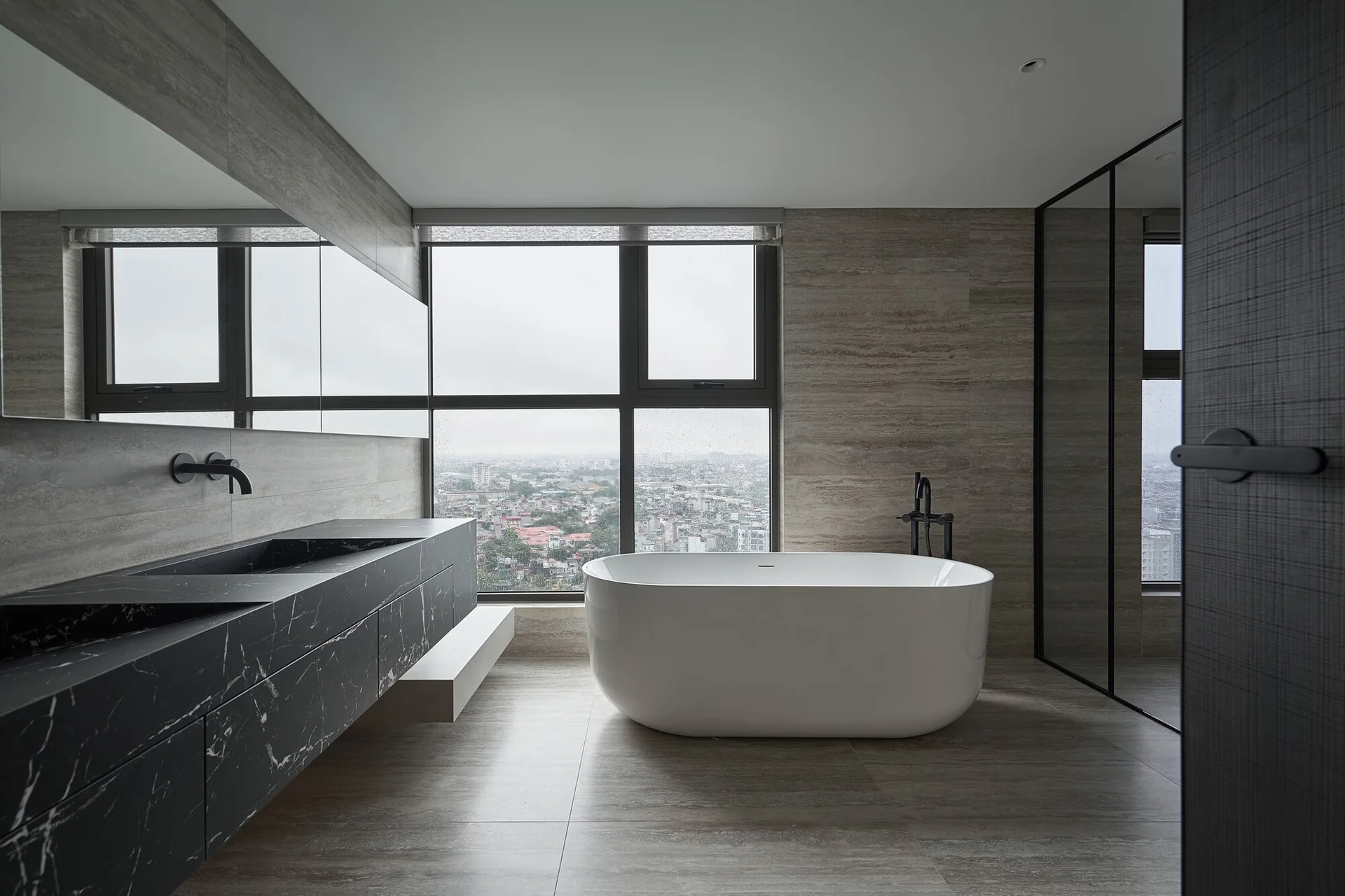 A minimalist gray bathroom features a white freestanding bathtub by a large window and a wall-mounted black marble vanity.