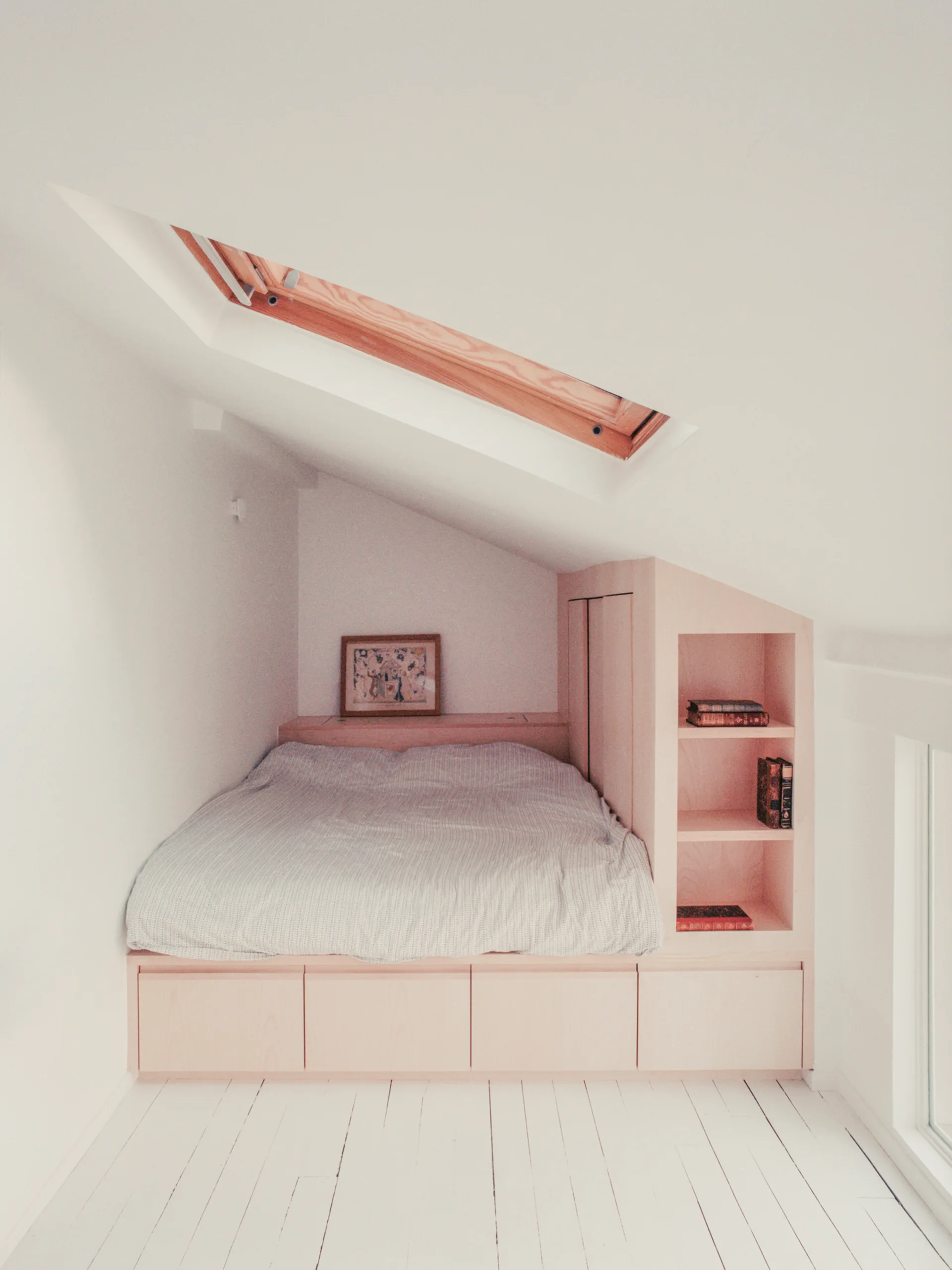 A light wood built-in platform bed with integrated shelving is tucked under a sloped white ceiling with a skylight and white wooden floors.