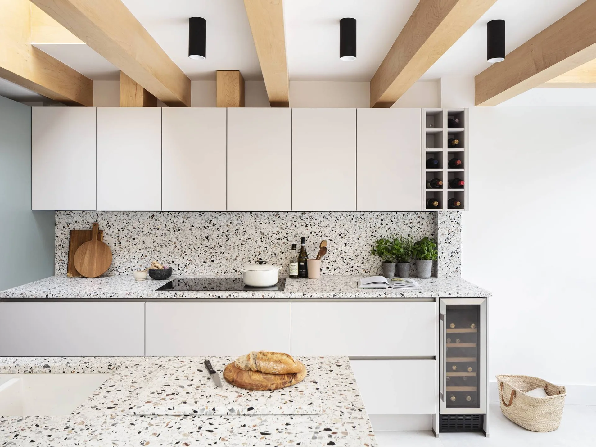 A modern white kitchen features speckled terrazzo countertops, a matching backsplash, flat-front cabinets, and exposed wood ceiling beams.