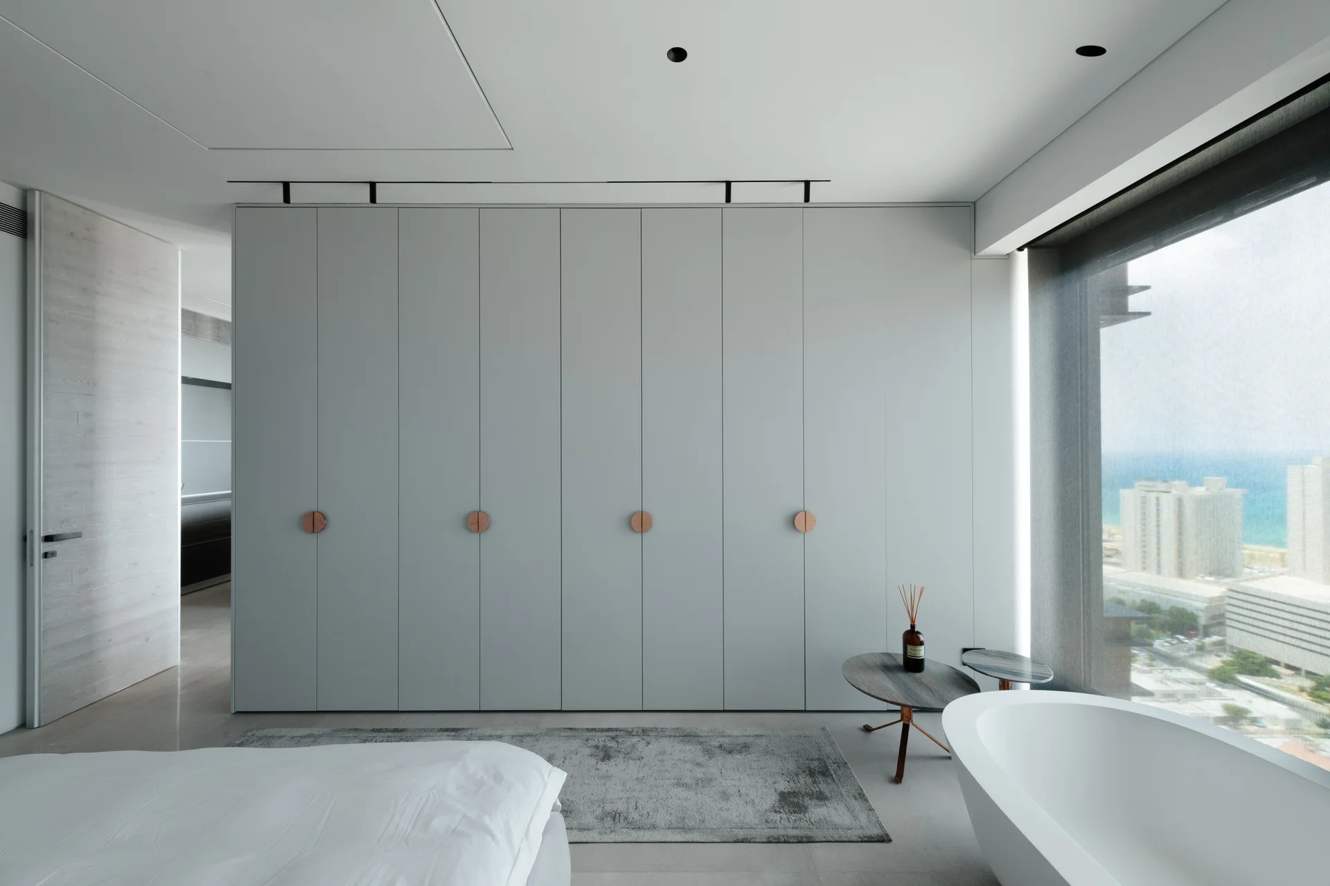 A minimalist bedroom features a large light grey floor to ceiling wardrobe with circular copper pulls, a freestanding white bathtub, and a window.