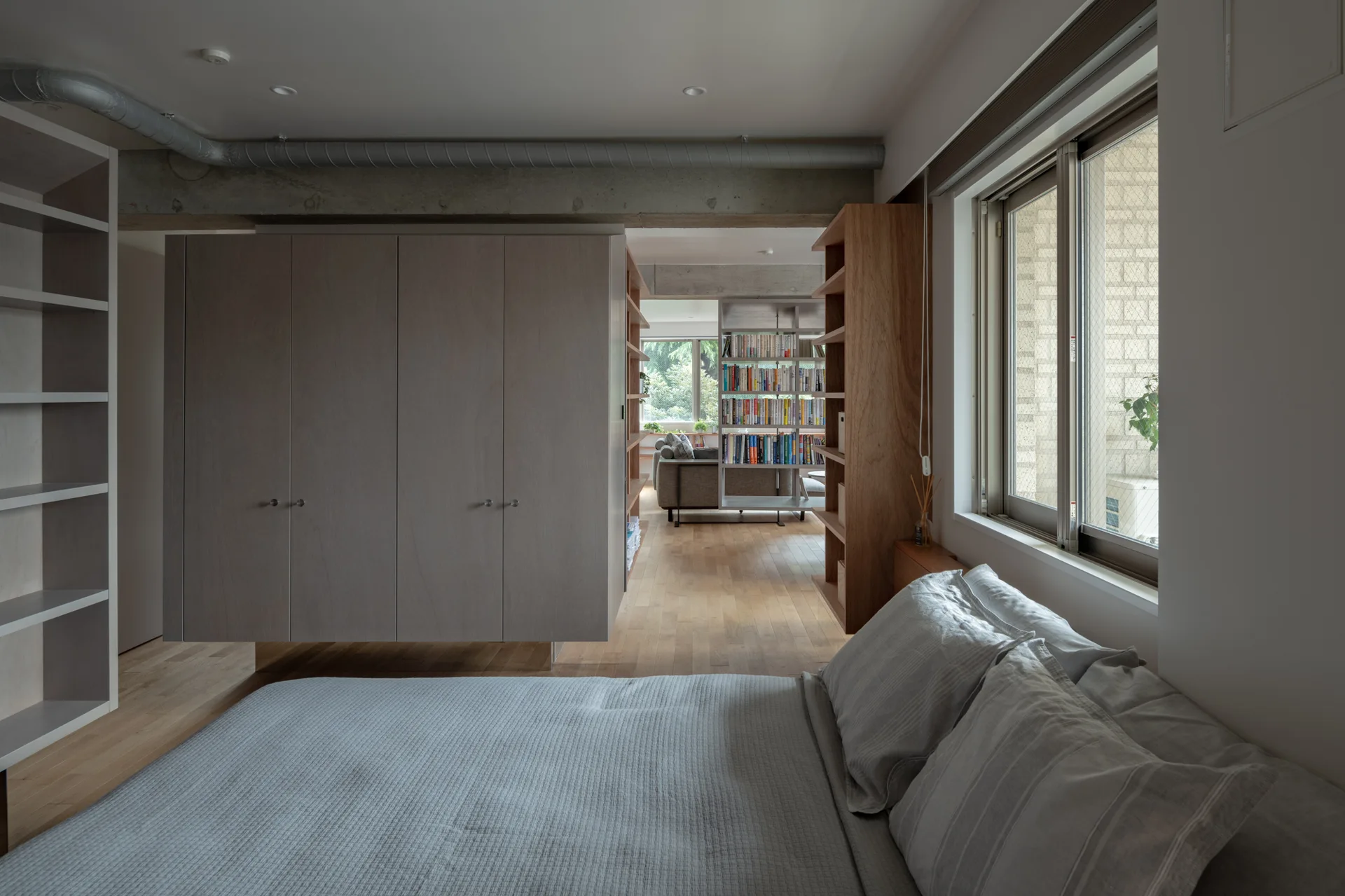 A grey bed sits next to a large suspended wooden wardrobe that divides the bedroom from an adjacent living area with bookshelves.