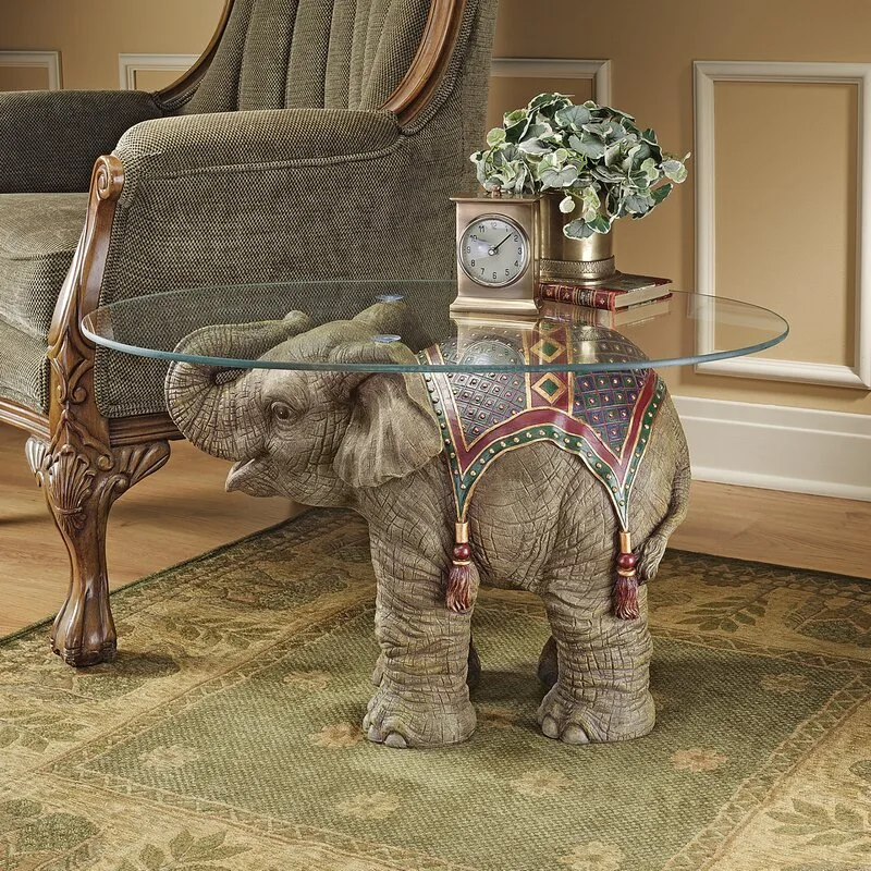 An elephant-shaped sculptural accent table with a round glass top sits on a green patterned rug beside a classic upholstered armchair.