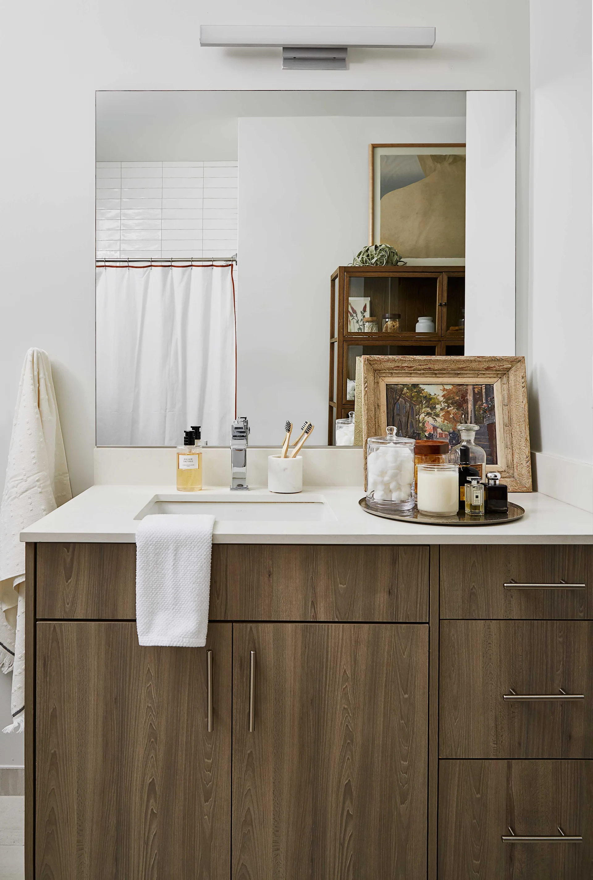 A wood bathroom vanity features a white countertop, chrome faucet, a large frameless mirror, and a tray of toiletries alongside a framed painting.
