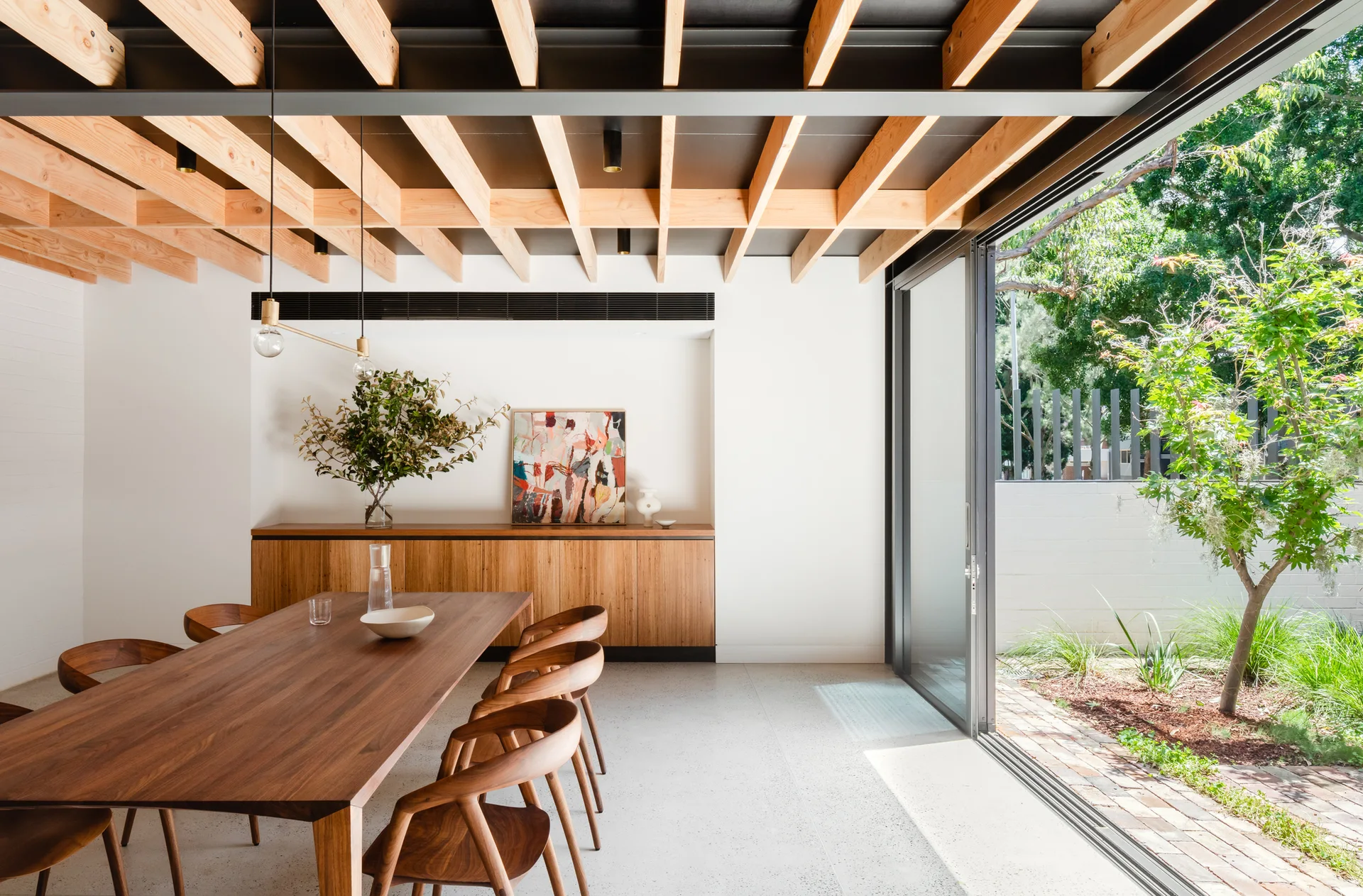 A dining room with a wooden table, matching chairs, and a low sideboard beneath an exposed timber joist ceiling next to sliding glass doors.