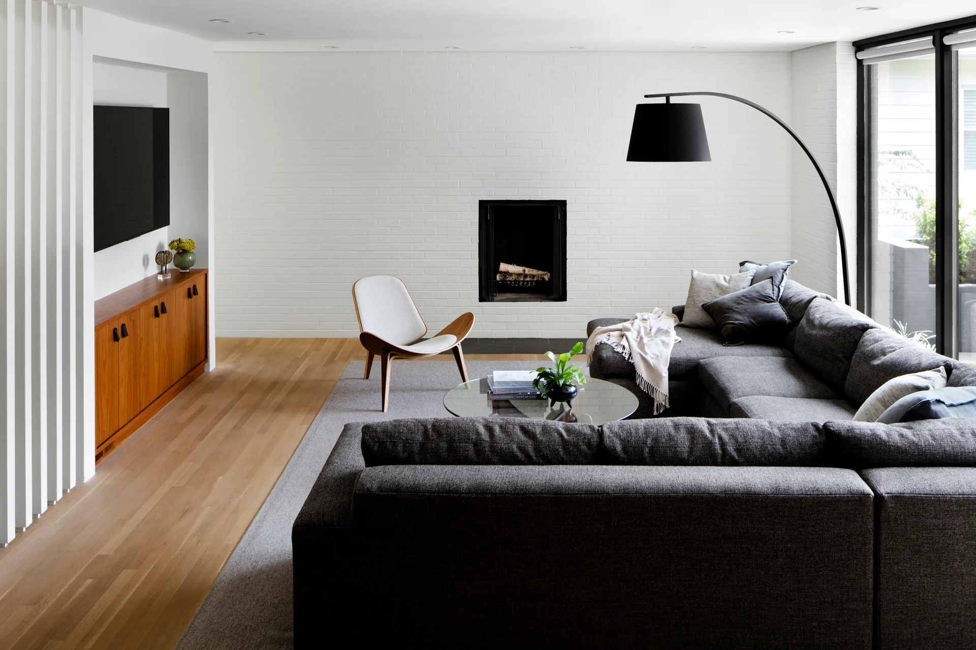 A minimalist living room features a charcoal sectional sofa, a white painted brick fireplace, and a wood media console.
